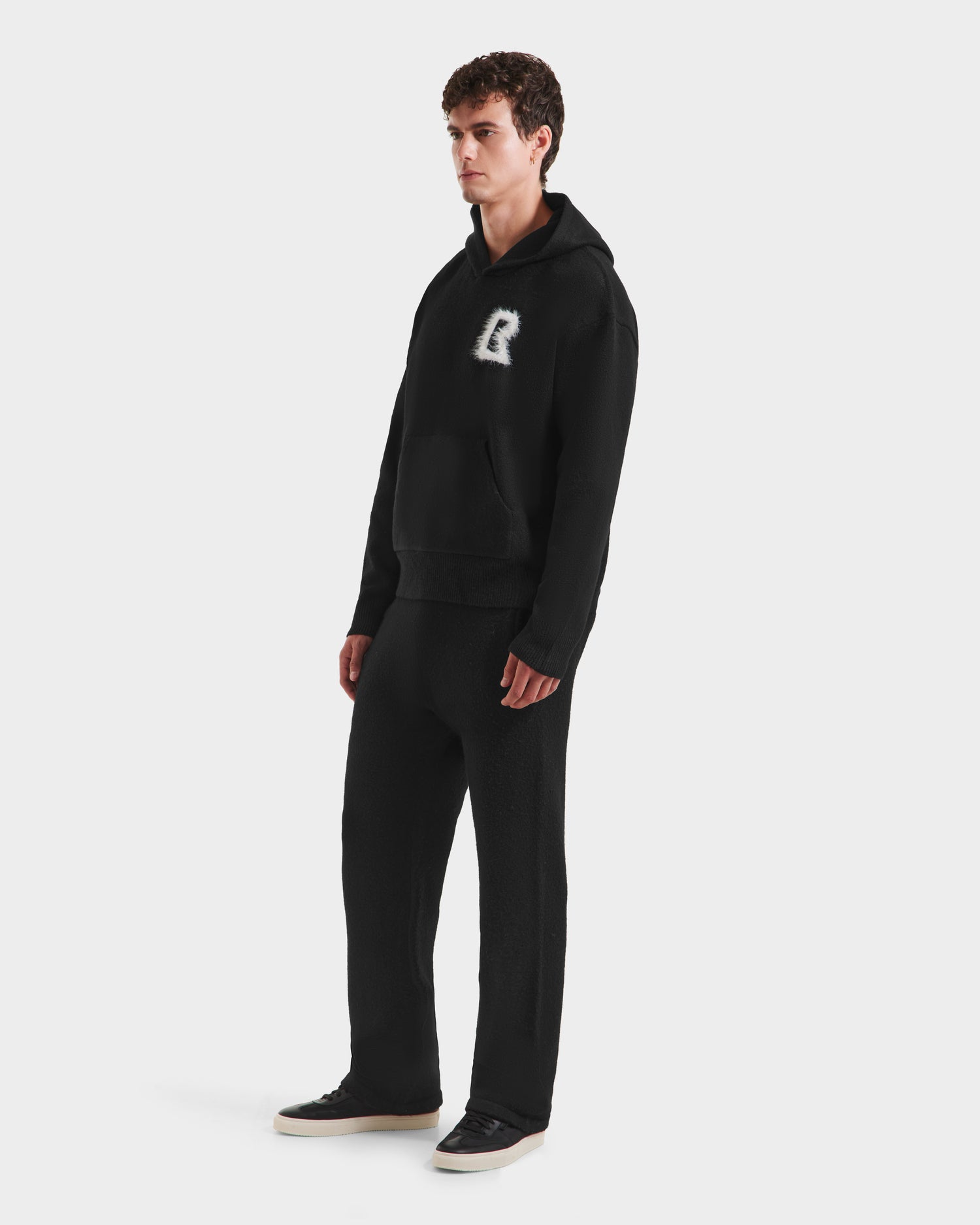 A man poses against a plain white background wearing Rudsak’s RAPHAEL - 6525643 Black hoodie featuring a fuzzy R logo, paired with matching black loungewear sweatpants and black sneakers with white soles.
