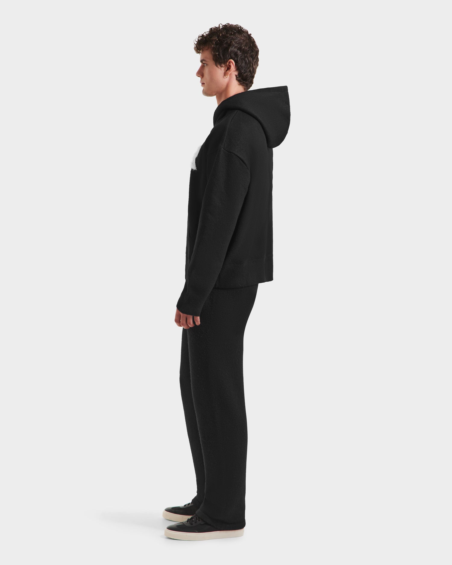 A person with short curly hair stands in profile, wearing the RAPHAEL - 6525643 Black hoodie by Rudsak and matching streetwear-inspired pants and sneakers with white soles, set against a plain white background.