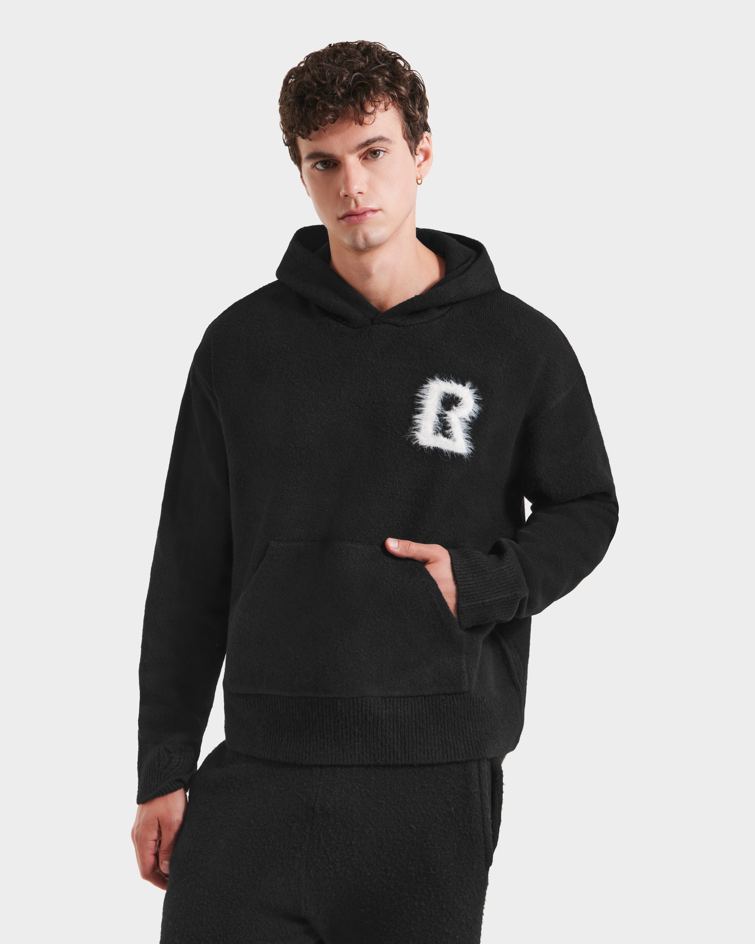 A young man models the RAPHAEL - 6525643 Black men’s hoodie by Rudsak, featuring a white fuzzy R logo and matching black loungewear pants, standing with one hand in the pocket against a light backdrop.