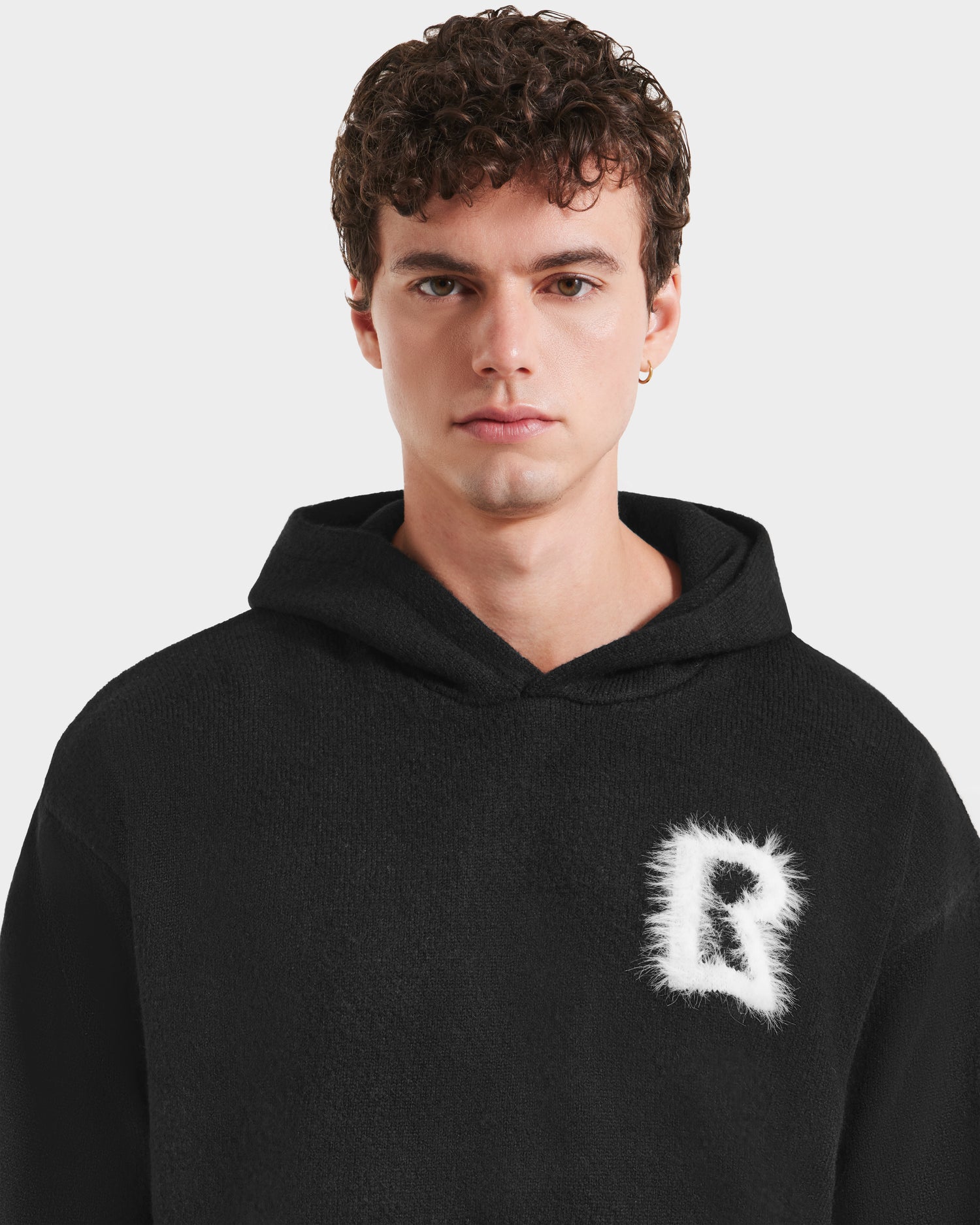 A young man with short curly hair models the RAPHAEL - 6525643 Black men’s hoodie by Rudsak, featuring a white, furry letter R on the left chest, styled against a light background.