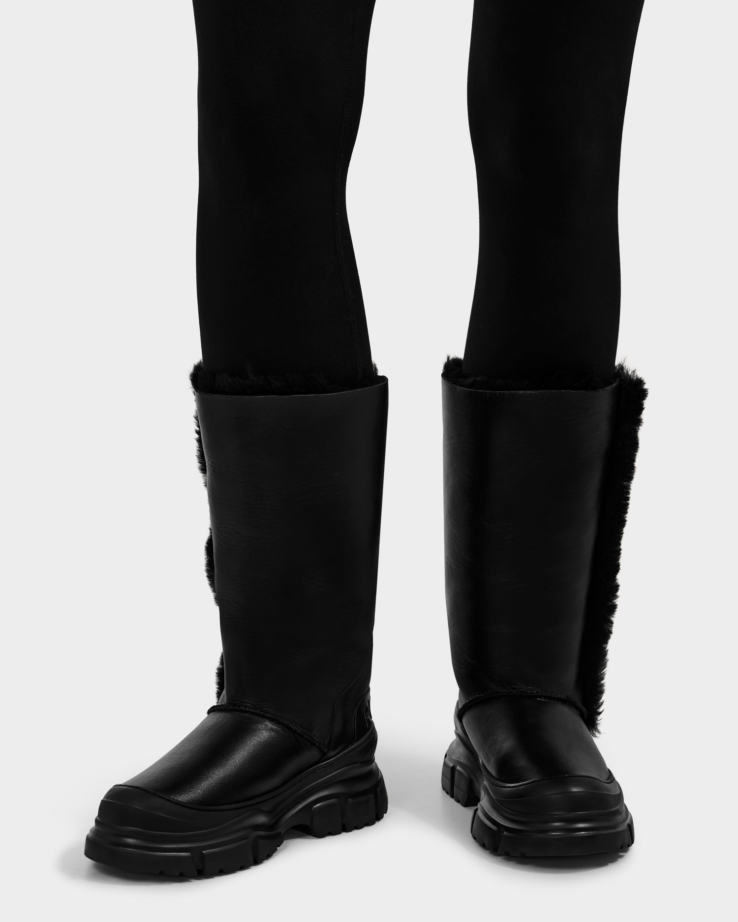 A person stands on a light gray background wearing Rudsak’s RAVENNA - 8225031 Black leather shearling knee-high boots with fur lining and black leggings—stylish, warm women’s winter boots.