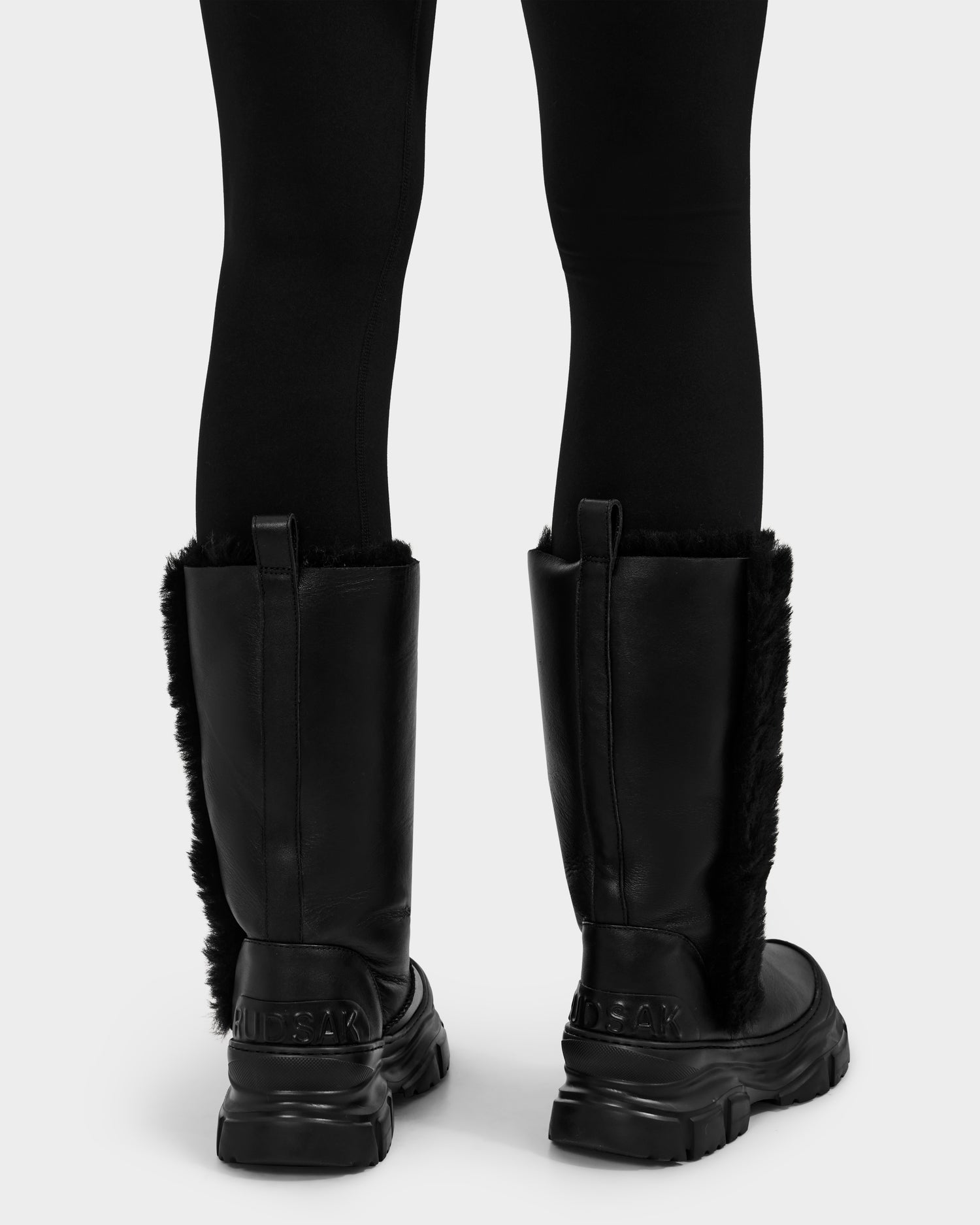A person wearing black leggings and Rudsak RAVENNA - 8225031 Black women's winter boots with faux fur lining and thick soles, viewed from the back against a white background.
