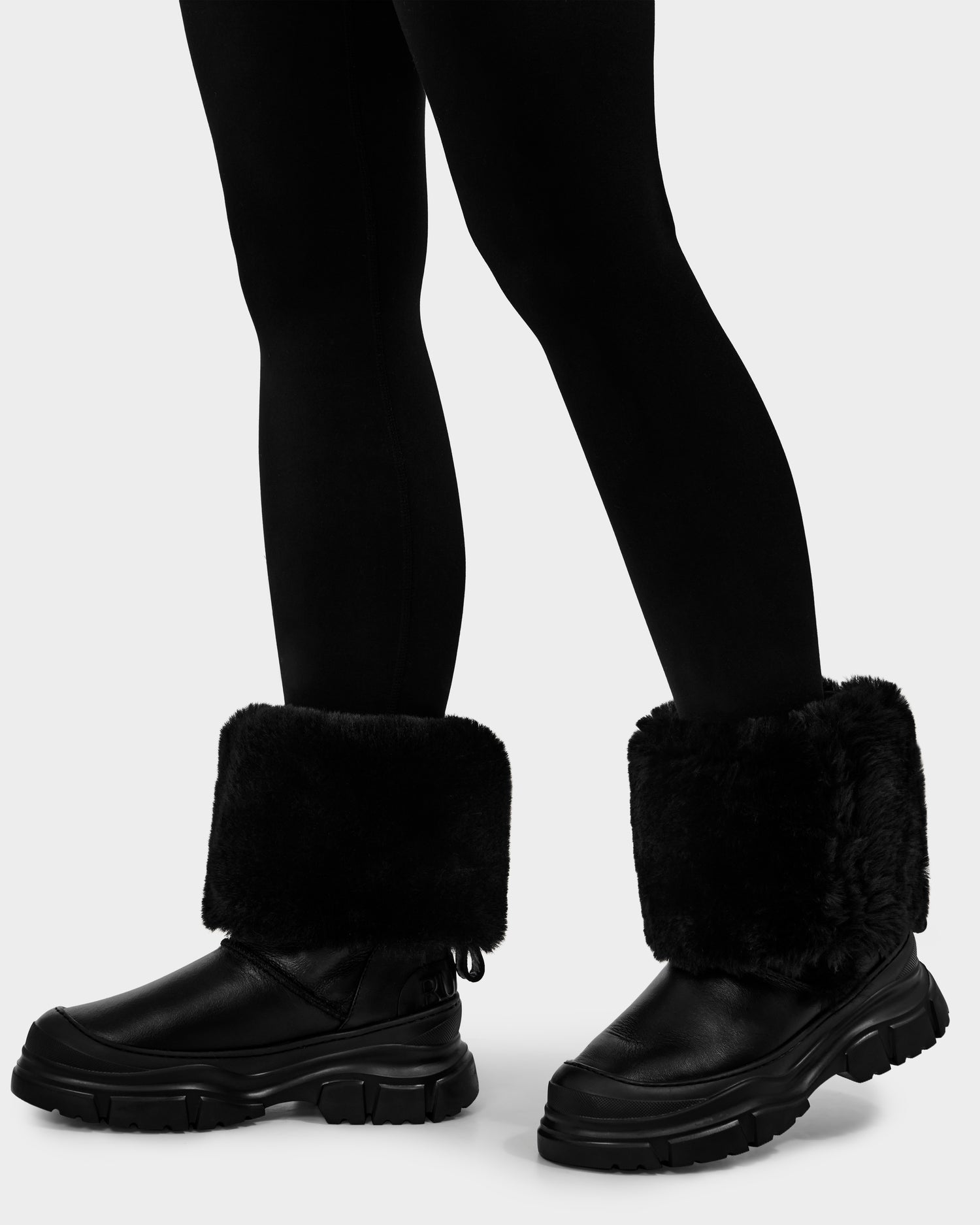 A person in black leggings and Rudsak RAVENNA - 8225031 Black women’s winter boots with thick faux fur cuffs and chunky rubber soles stands against a plain light background.