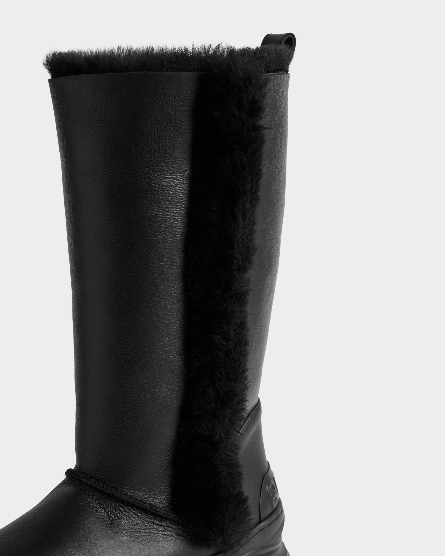 A close-up of the Rudsak RAVENNA - 8225031 Black women’s high winter boot showcases its textured leather exterior, vertical fur trim, and top loop, all set against a plain light gray background.