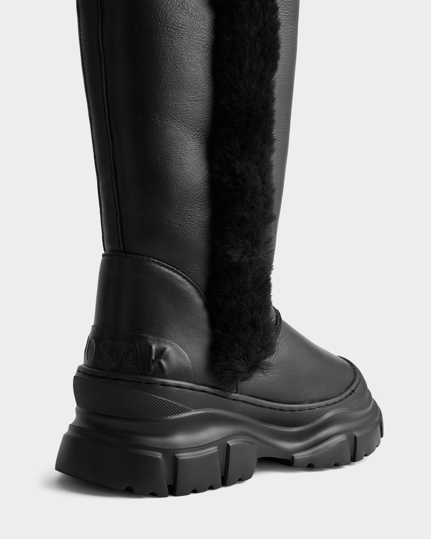 A close-up of the Rudsak RAVENNA - 8225031 Black women’s high winter boot with a black leather upper, chunky sole, black shearling trim on the side, and embossed branding near the heel, shown against a plain light background.