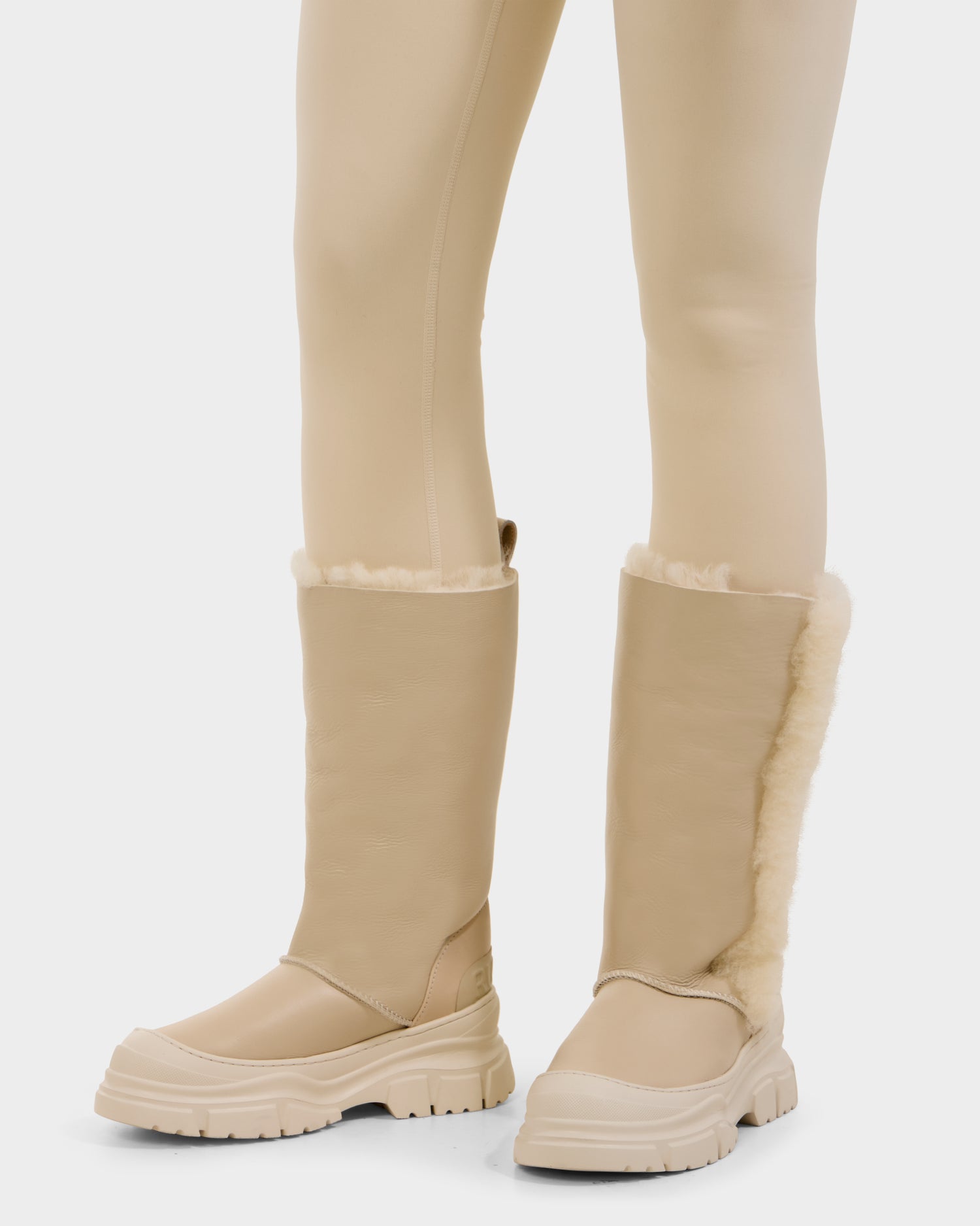 A person wears the Rudsak RAVENNA - 8225031 Vanilla women’s high winter boots—tan, knee-high leather shearling with chunky rubber soles and exposed trim—paired with beige leggings against a plain white background.