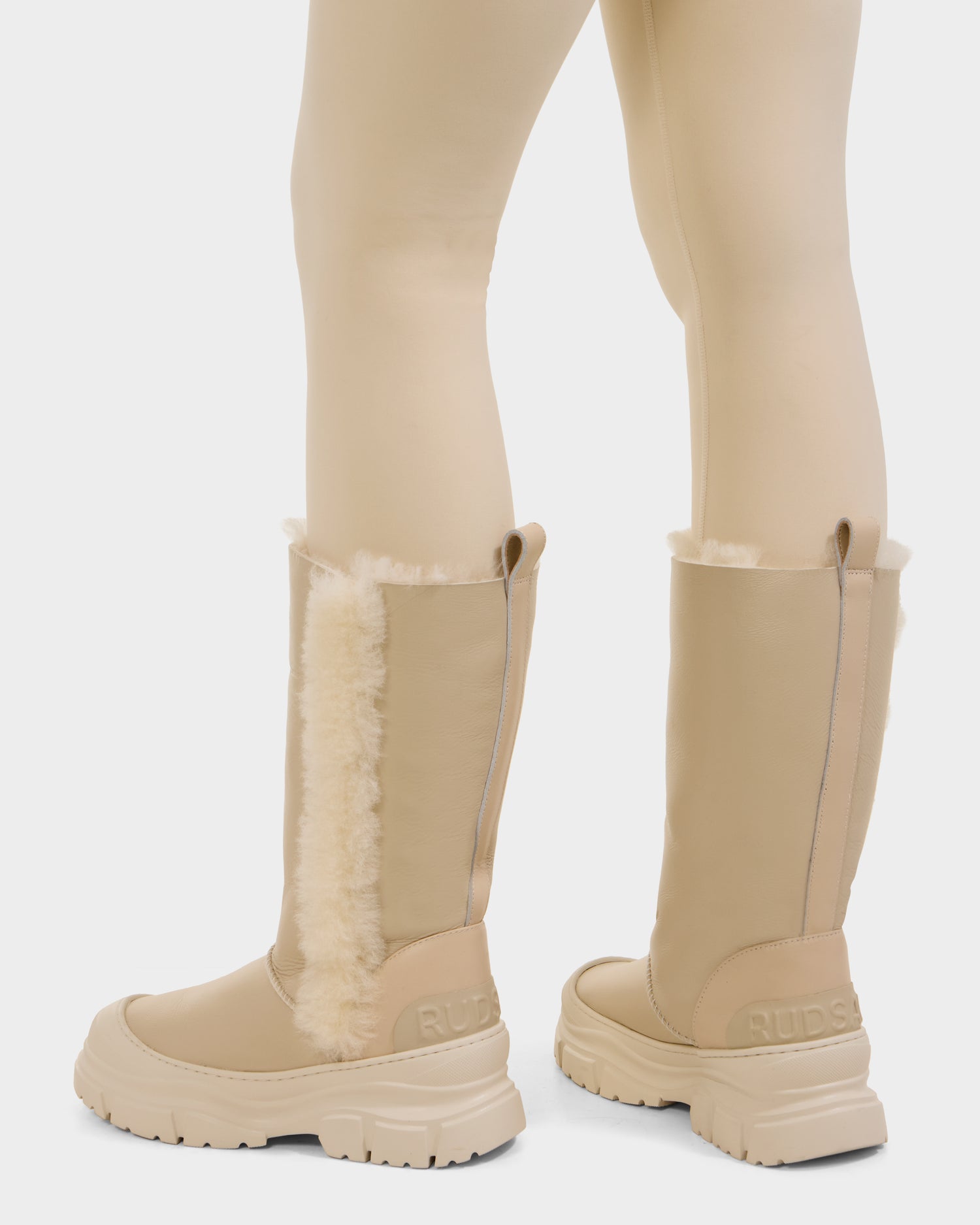 A person is shown from the back wearing Rudsak's RAVENNA - 8225031 Vanilla women's winter boots with chunky soles and cream faux fur trim, paired with beige leggings. These warm, stylish boots are perfect for cozy everyday wear.