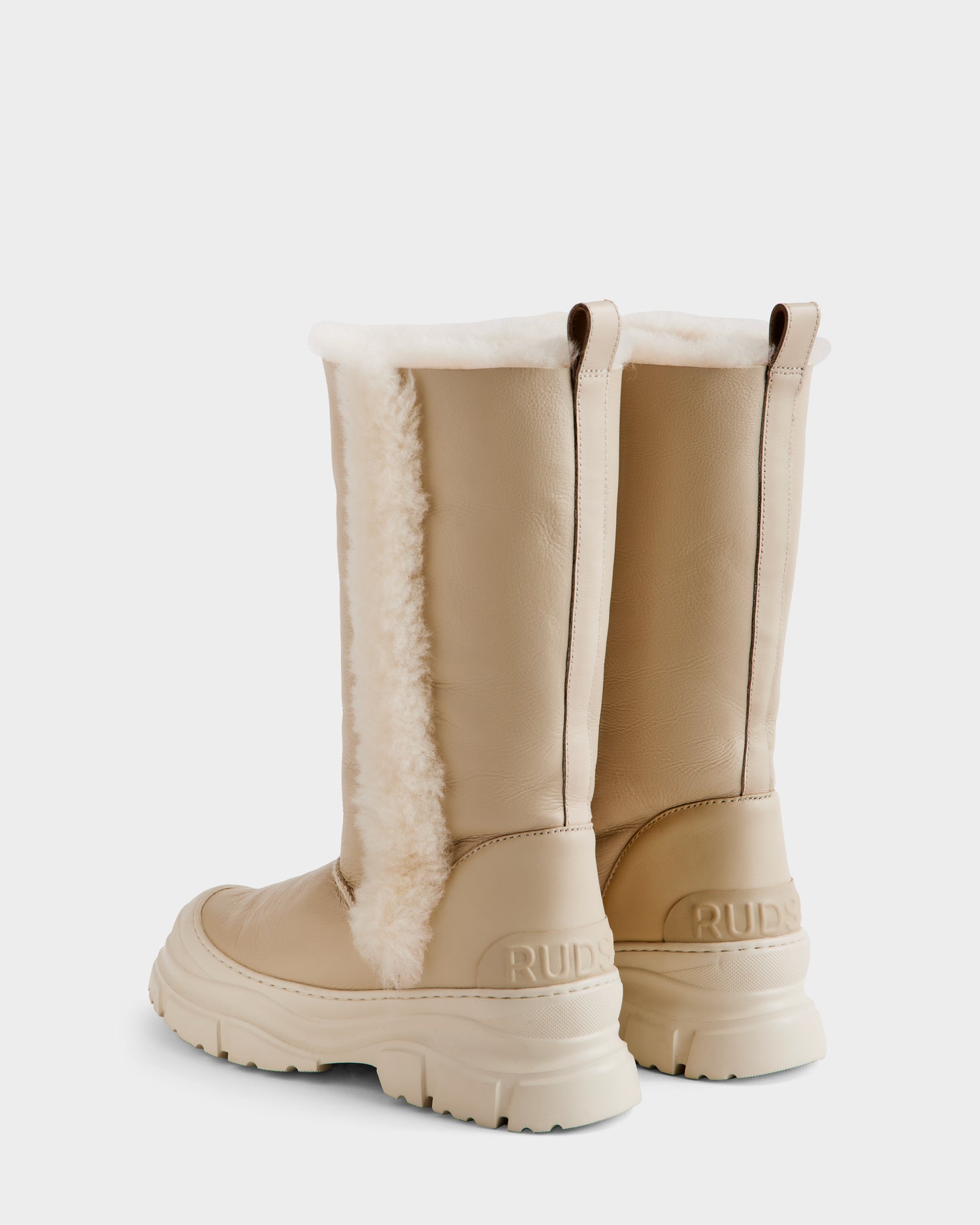 The RAVENNA - 8225031 Vanilla by Rudsak is a women's knee-high winter boot in beige, featuring chunky rubber soles, white faux shearling lining, pull tabs on top, and embossed branding near the heels, shown from the back and side.