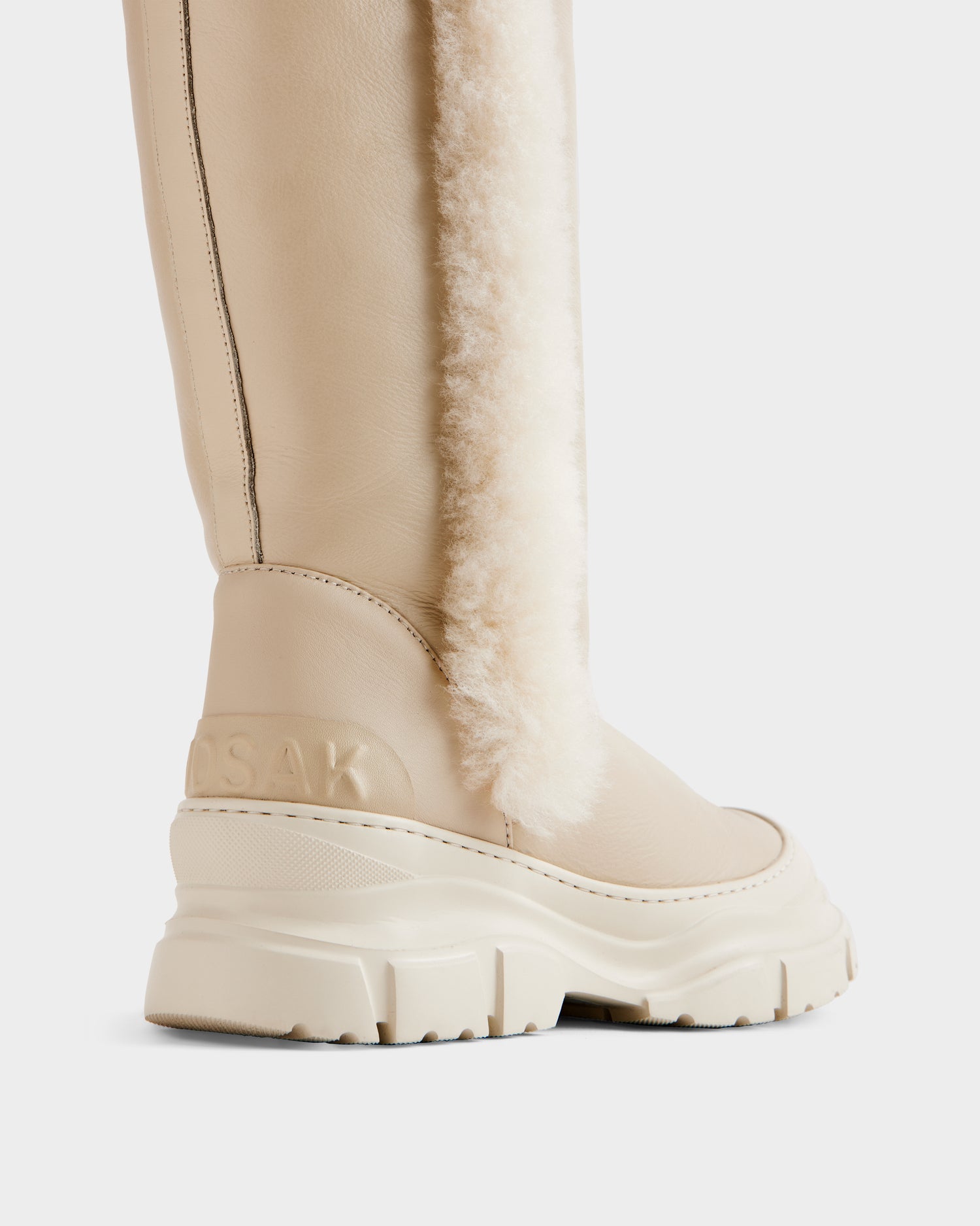 Close-up of the RAVENNA - 8225031 Vanilla women’s high winter boot by Rudsak, featuring a beige leather exterior, chunky sole, vertical white shearling strip, stitched back detailing, and embossed branding on the heel.