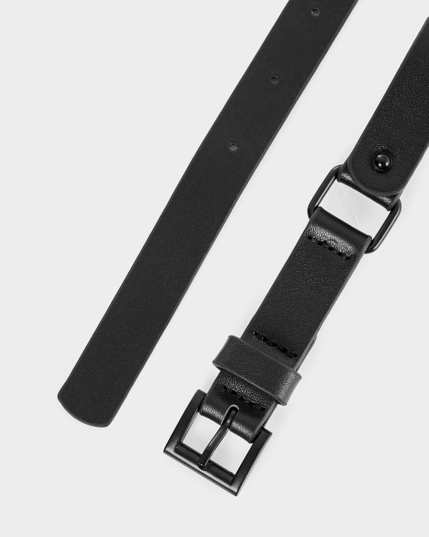 A close-up of the Rudsak RAYNA - 8425539 Black leather harness belt with a black rectangular metal buckle on a white background. The belt features multiple holes for waist cinching and a smooth, matte finish.