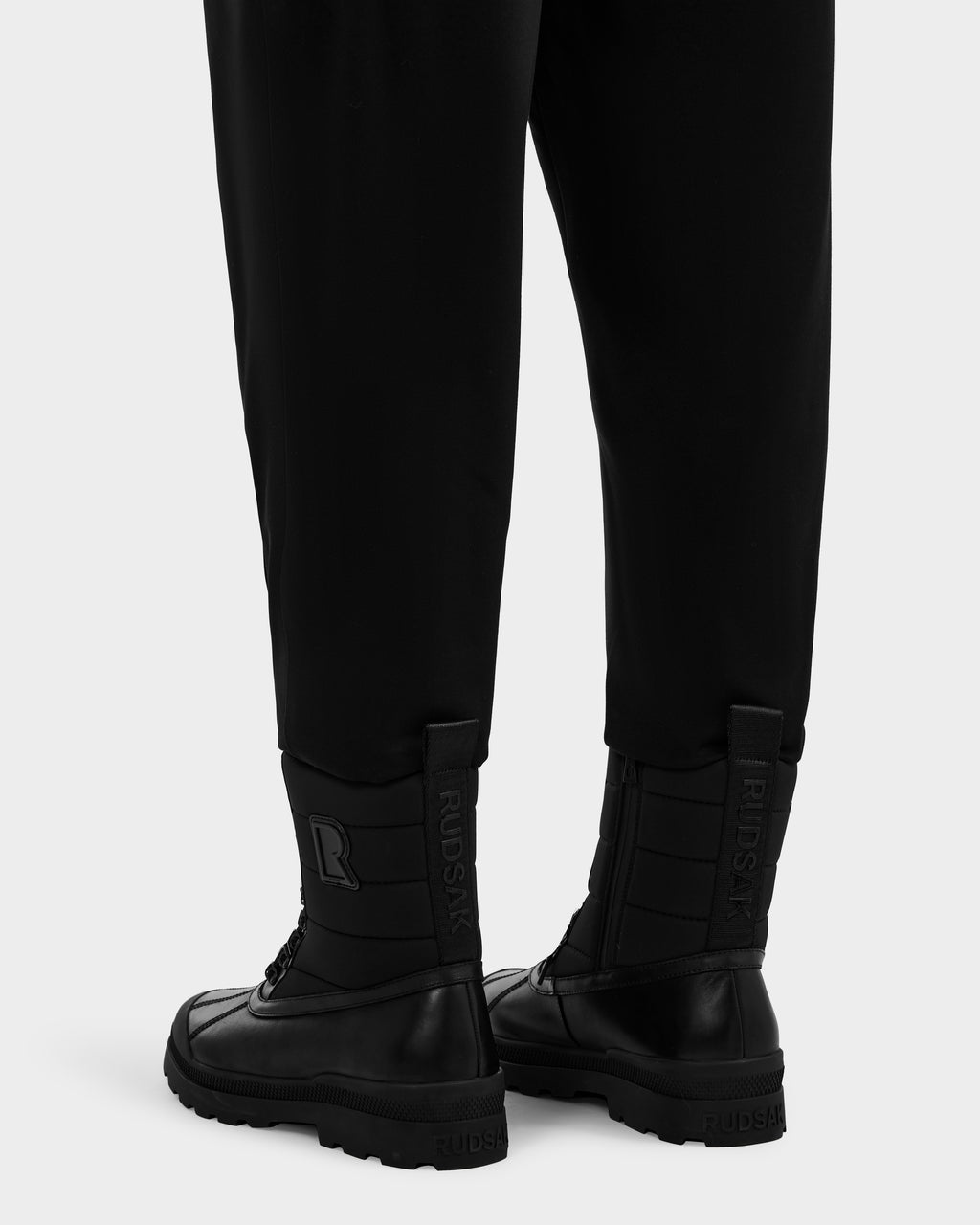 A person wears black pants and Rudsak REAGAN - 6224501 Black leather boots with thick soles and high tops, featuring Rudsak branding and a large R embossed on the heel tabs. Photo is shot from behind at ankle level.