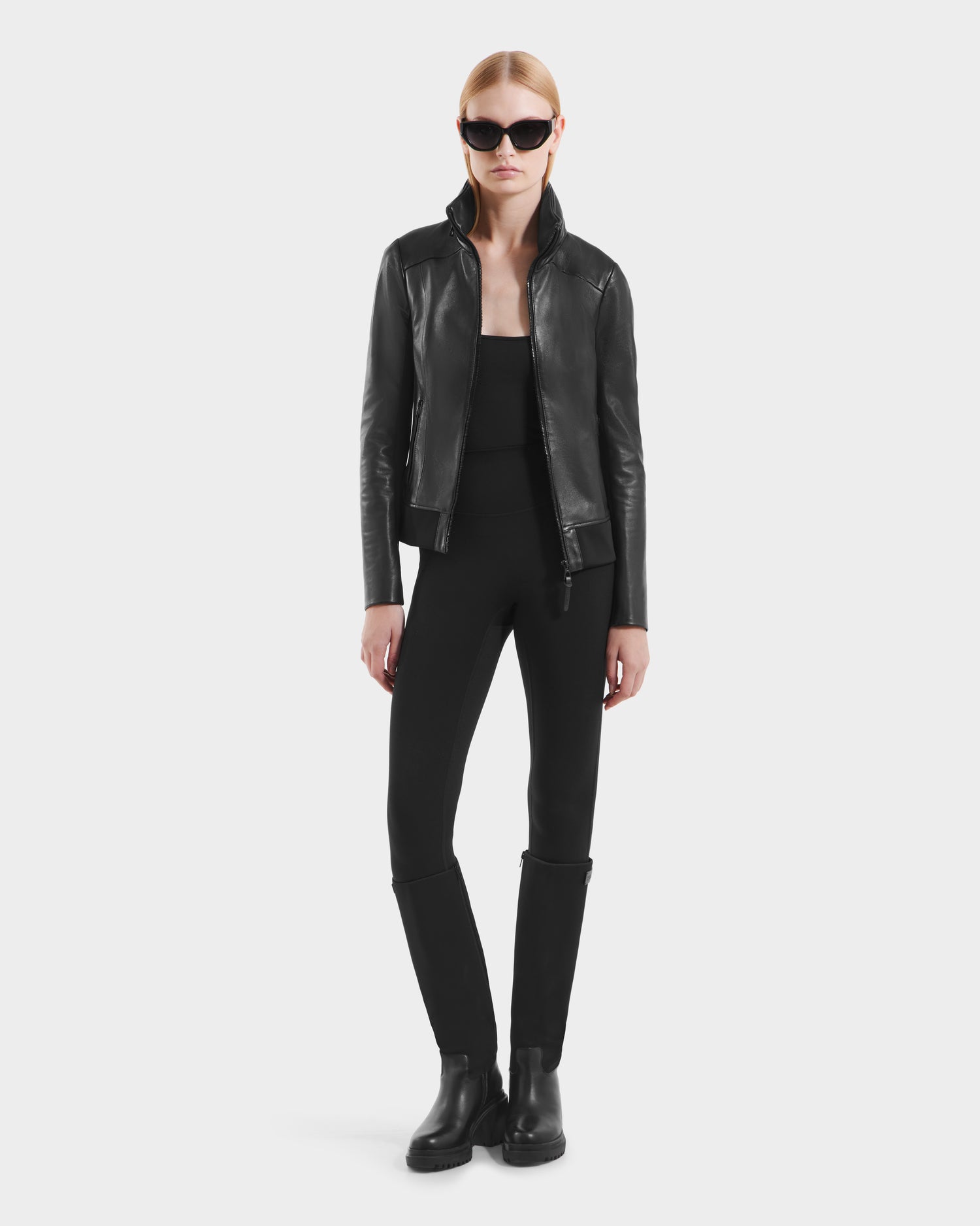 A woman in Rudsaks REBECCA - 8125066 Black lamb leather moto-inspired bomber with a lightweight hideaway hood, sunglasses, black top, fitted pants, and boots stands against a plain light background.