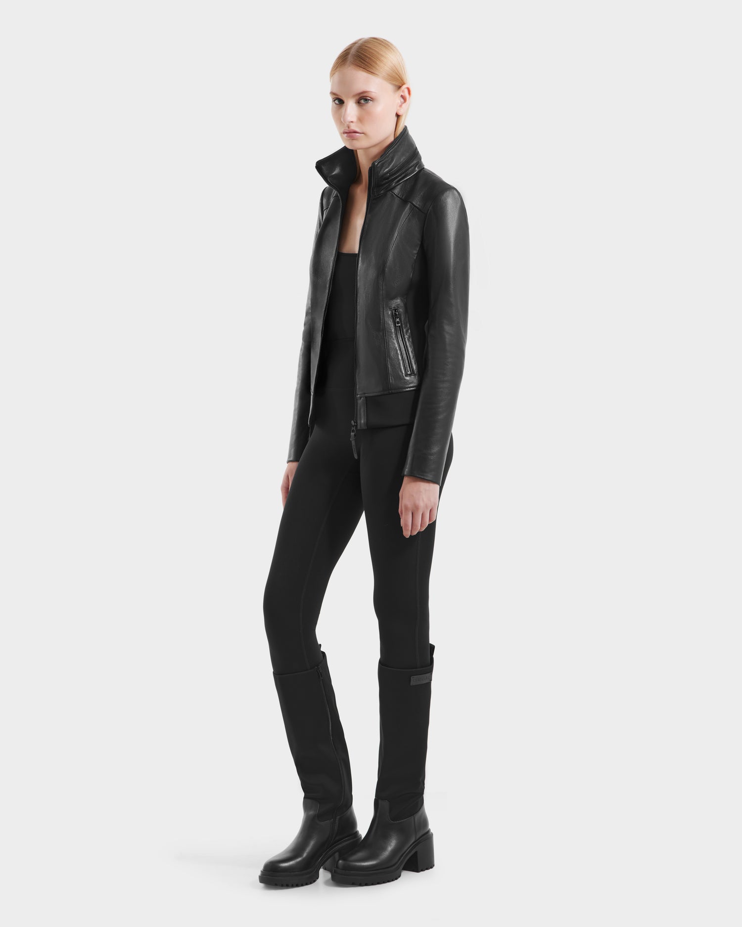 A person with blonde hair stands against a white background wearing the Rudsak REBECCA - 8125066 Black moto-inspired leather bomber jacket, paired with black pants and boots.