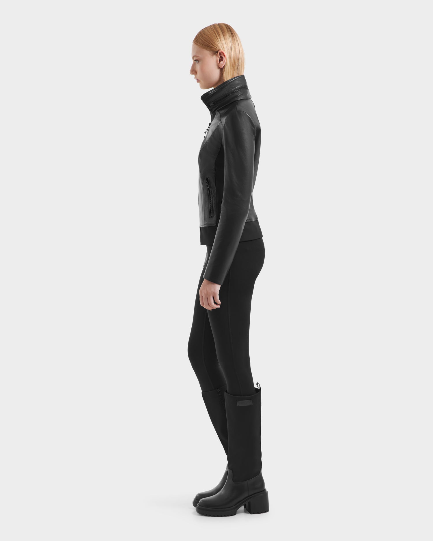 A woman wears the Rudsak REBECCA - 8125066 Black moto-inspired bomber, crafted from lamb leather, paired stylishly with black boots.