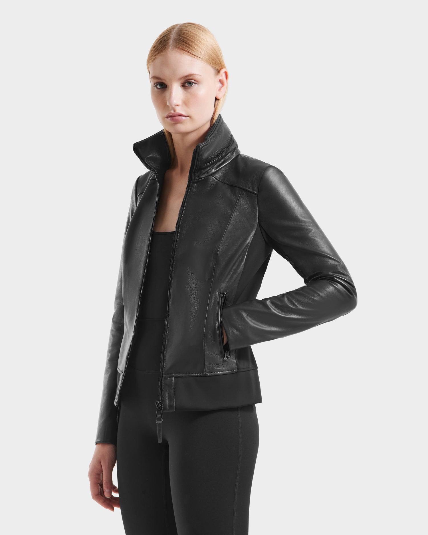 A woman with straight blonde hair wears the Rudsak REBECCA - 8125066 Black fitted lamb leather jacket over a black top and pants, standing against a plain light gray background.