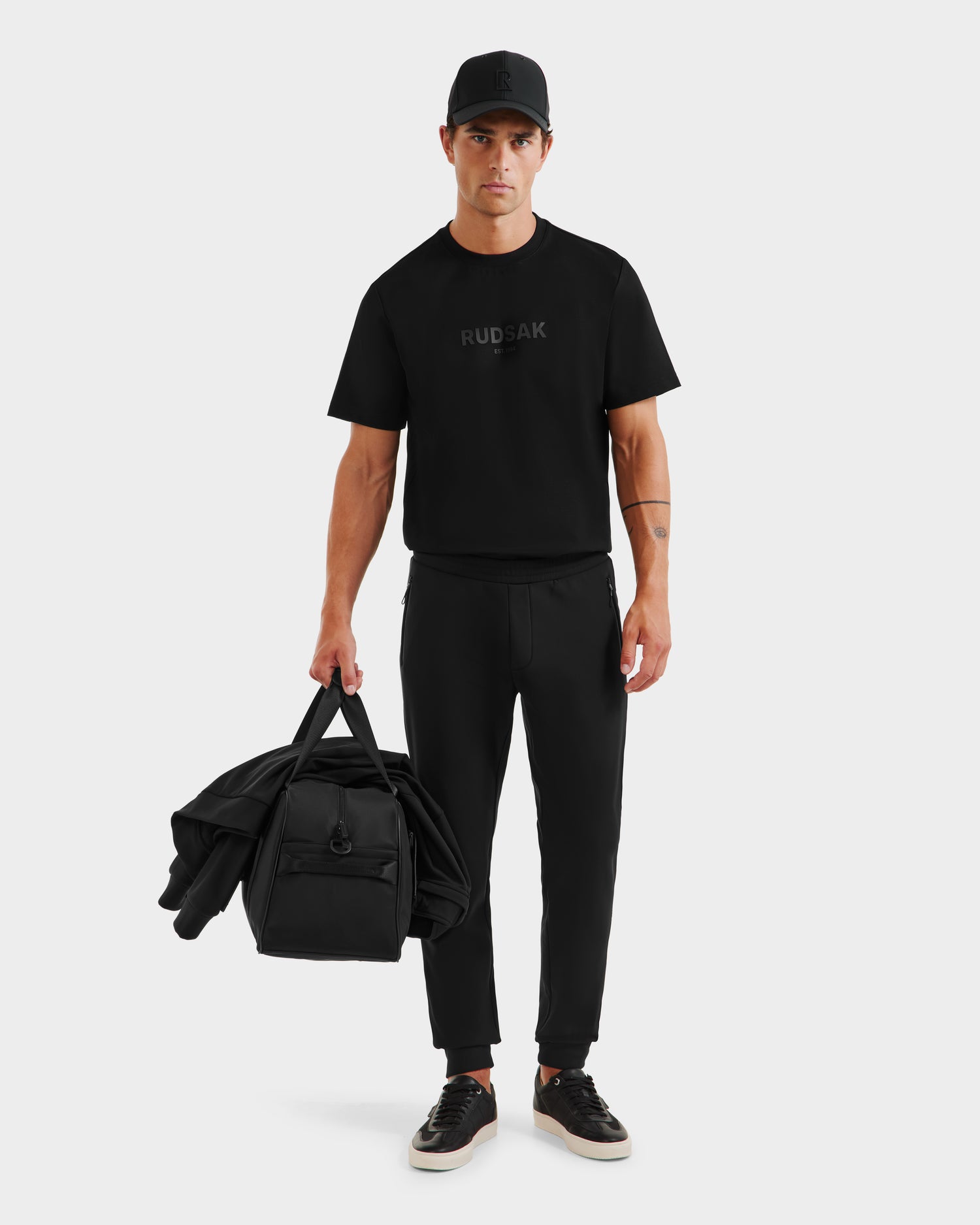 A man dressed in black Rudsak REGIS - 6525582 casual wear stands against a plain light background, holding a matching jacket and bag in his left hand.
