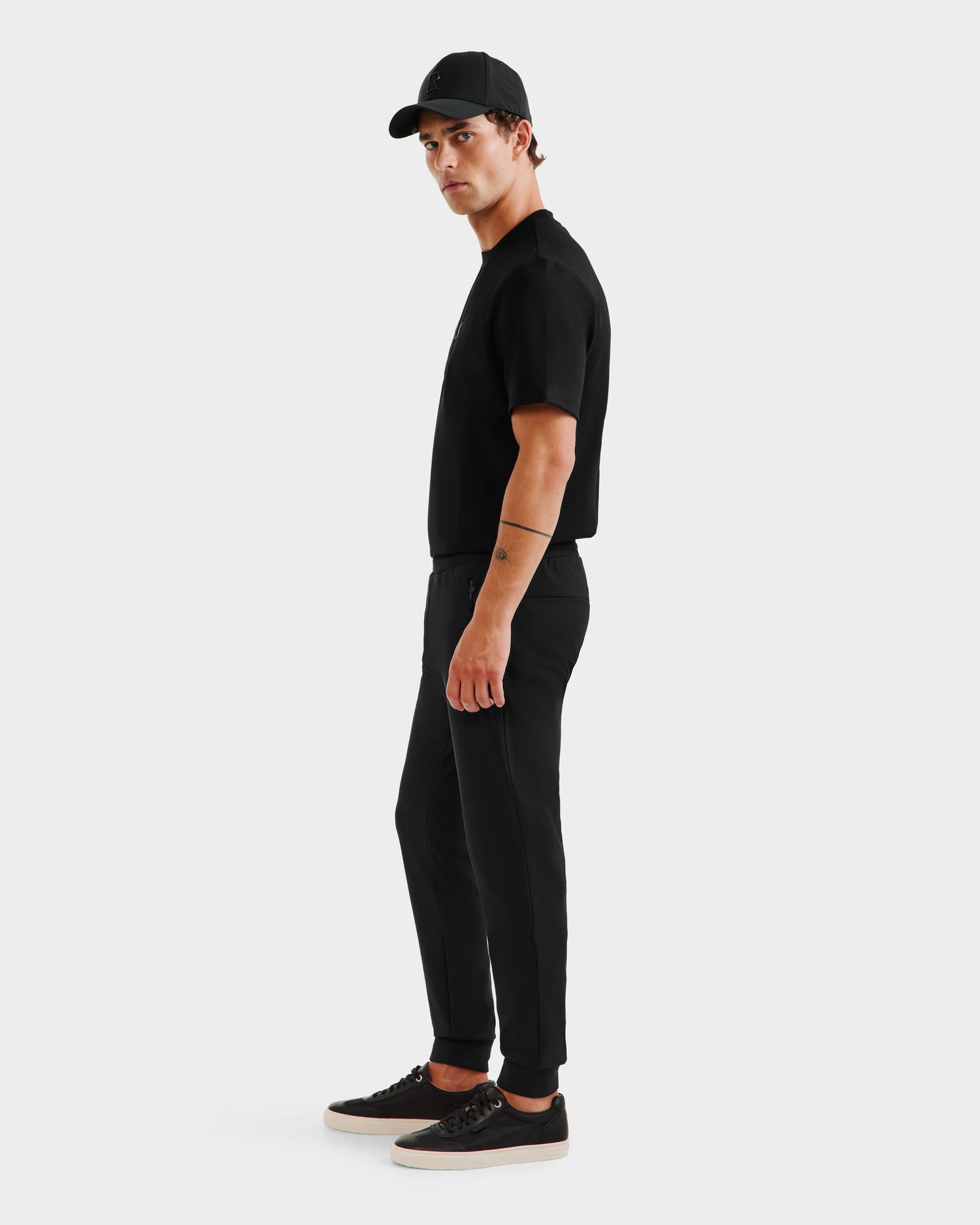 A man wearing Rudsaks REGIS - 6525582 Black men’s performance jogger pants, a black cap, and black t-shirt with black sneakers stands sideways against a white background, looking at the camera.