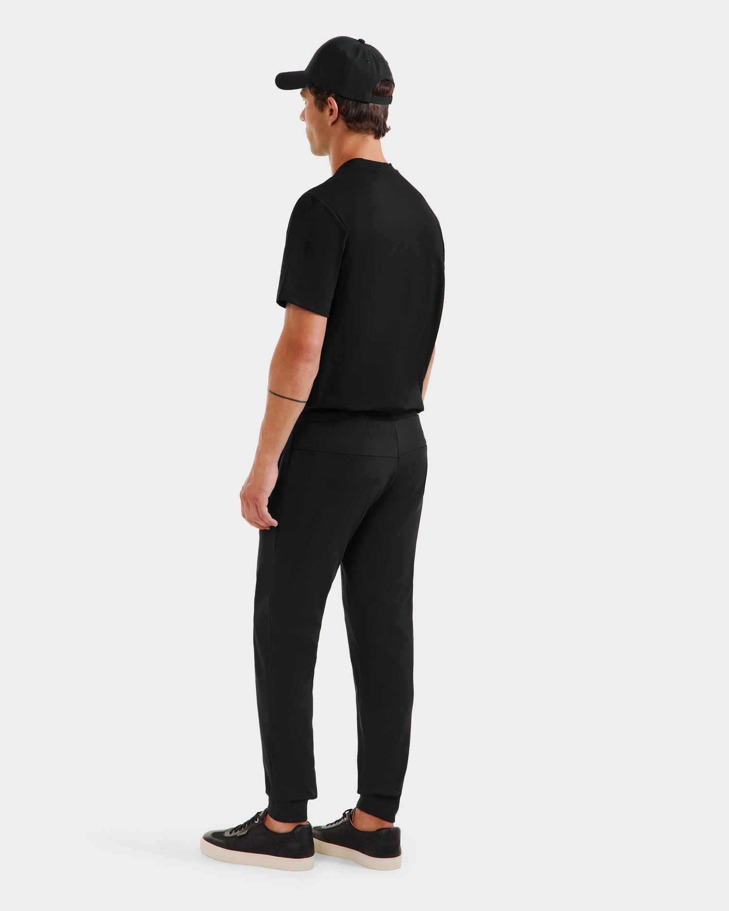 A man faces away, wearing the REGIS - 6525582 Black short-sleeve shirt from Rudsak, paired with stretch joggers, black sneakers with white soles, and a black cap against a plain light background.