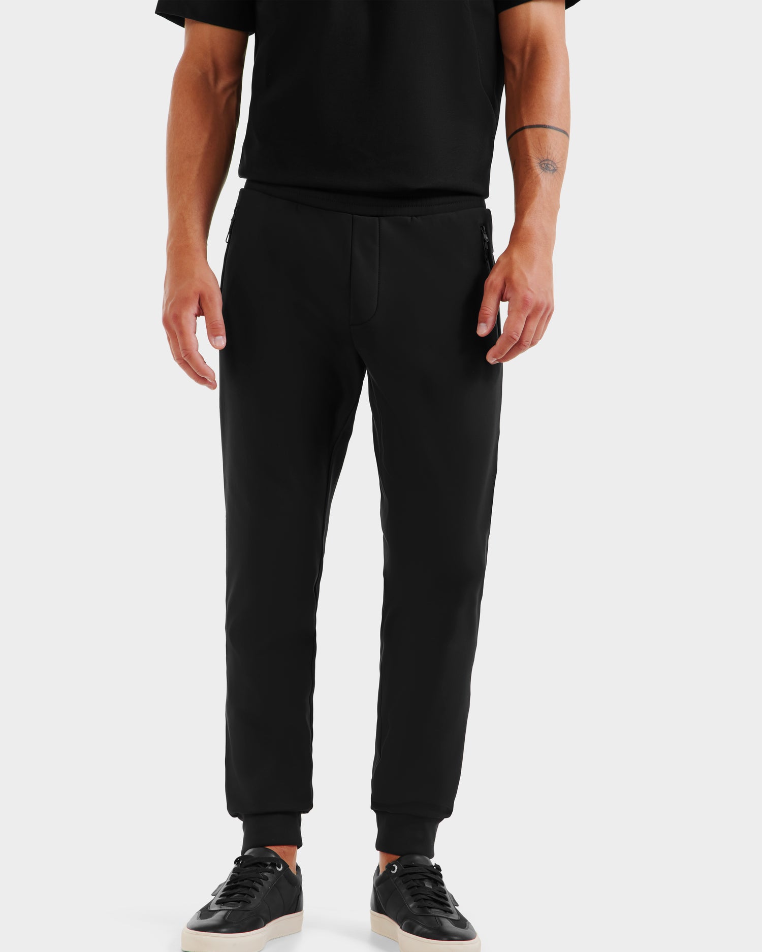 Wearing the REGIS - 6525582 Black short-sleeve shirt by Rudsak, men's stretch joggers, and black sneakers with white soles, a person stands against a white background, shown from shoulders to feet.