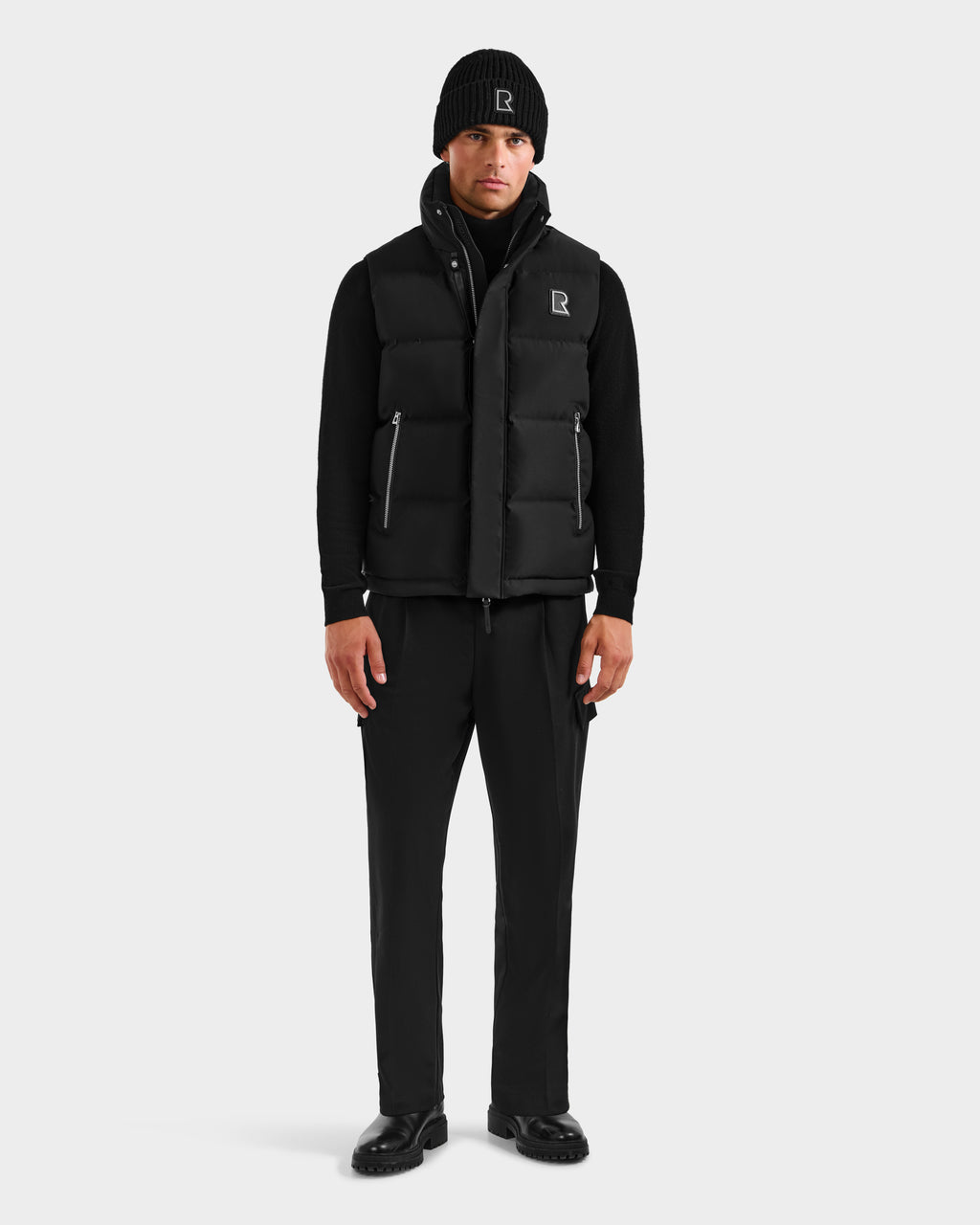 A person stands facing forward wearing the Rudsak REN H - 6125688 Black men's quilted down puffer vest with a removable hood, black beanie, long-sleeve shirt, pants, and shoes against a plain white background.