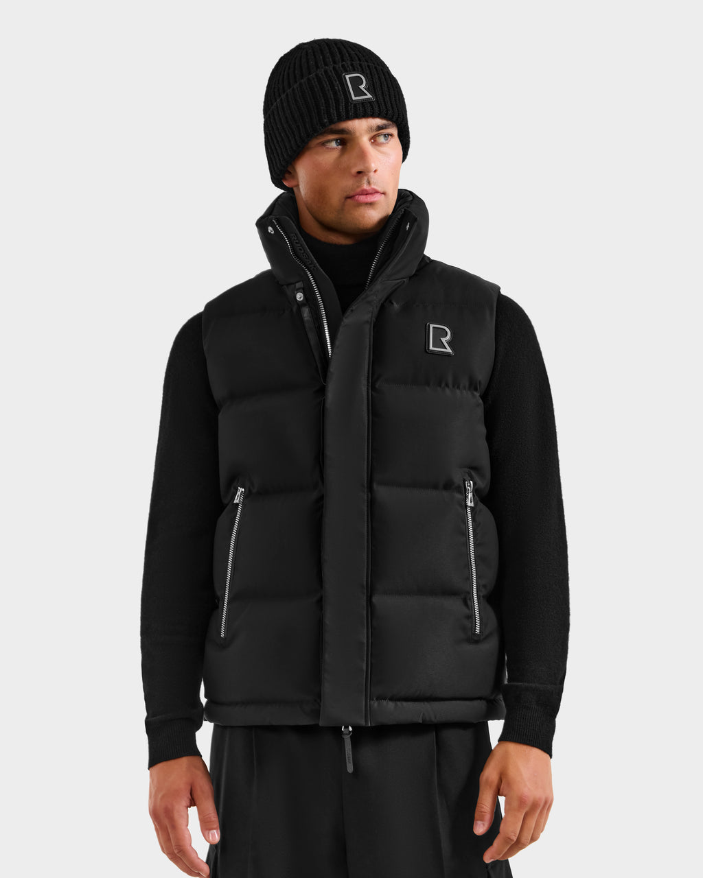 Wearing the Rudsak REN H - 6125688 Black men's quilted down puffer vest with zip and logo, paired with a black beanie, turtleneck, and pants—an essential cold-weather look against a light gray background.