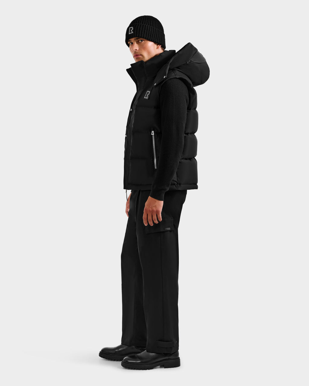 A man in a minimal, modern look models the Rudsak REN H - 6125688 Black men’s quilted down puffer vest with a removable hood, paired with a black long-sleeve shirt, pants, boots, and beanie against a plain light background.
