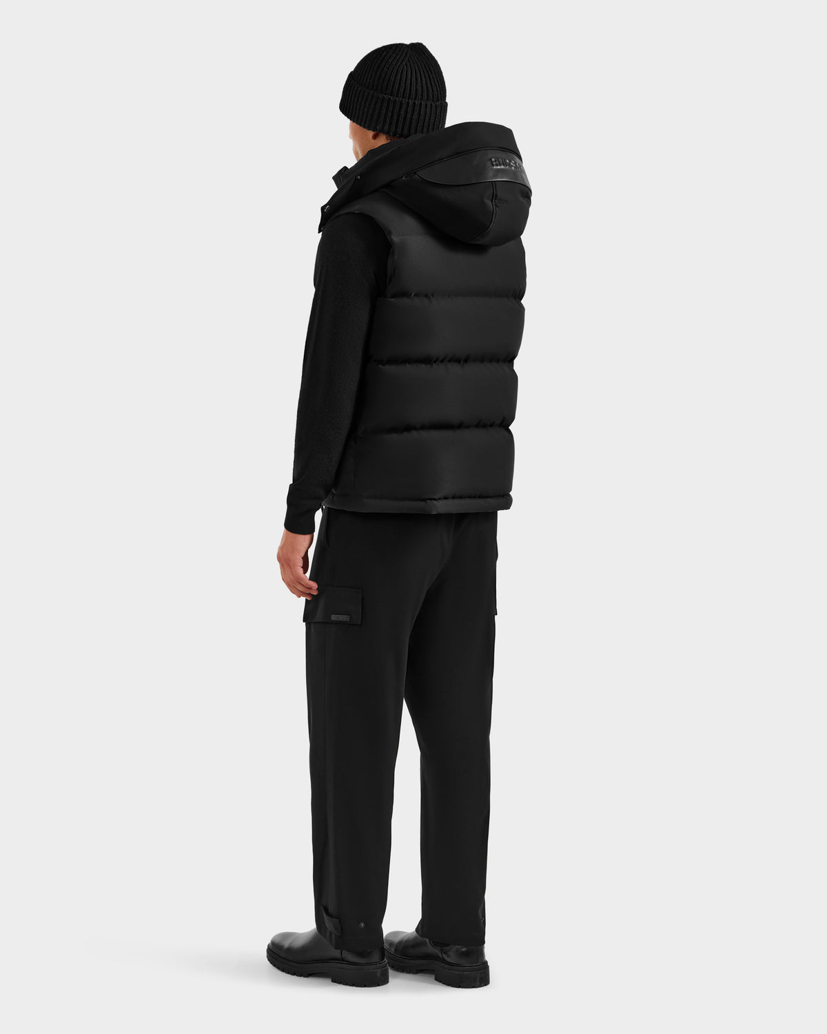 A person faces away wearing Rudsak’s REN H - 6125688 Black men's quilted down puffer vest with removable hood, paired with a black beanie, long-sleeve shirt, cargo pants, and shoes—ideal cold-weather essentials against a plain background.
