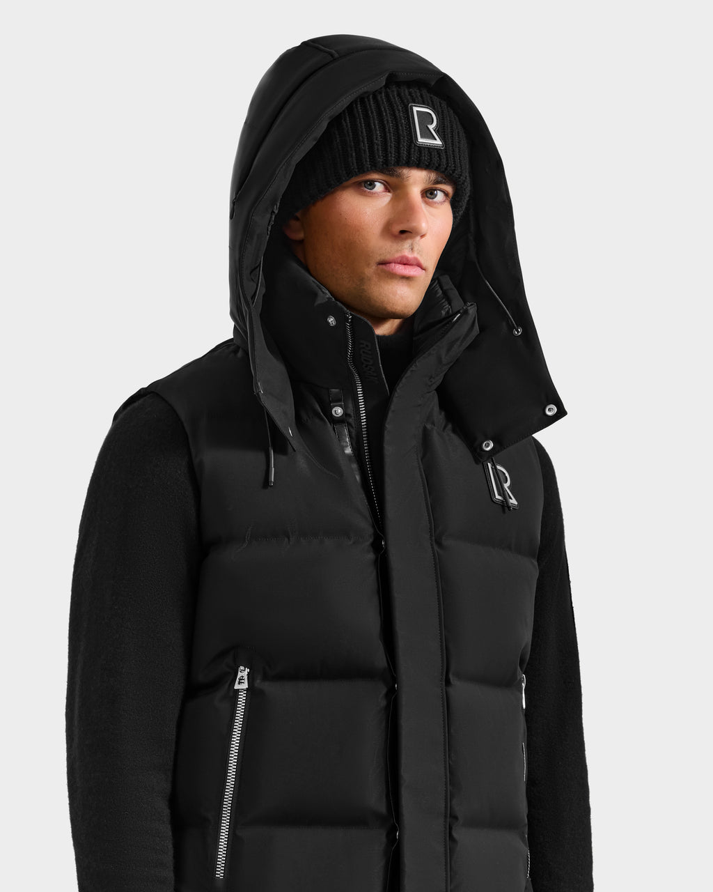 A man wearing the Rudsak REN H - 6125688 Black men's quilted down puffer vest with a removable hood, styled over a black sweater and R logo beanie, stands against a light background—a cold-weather essential.