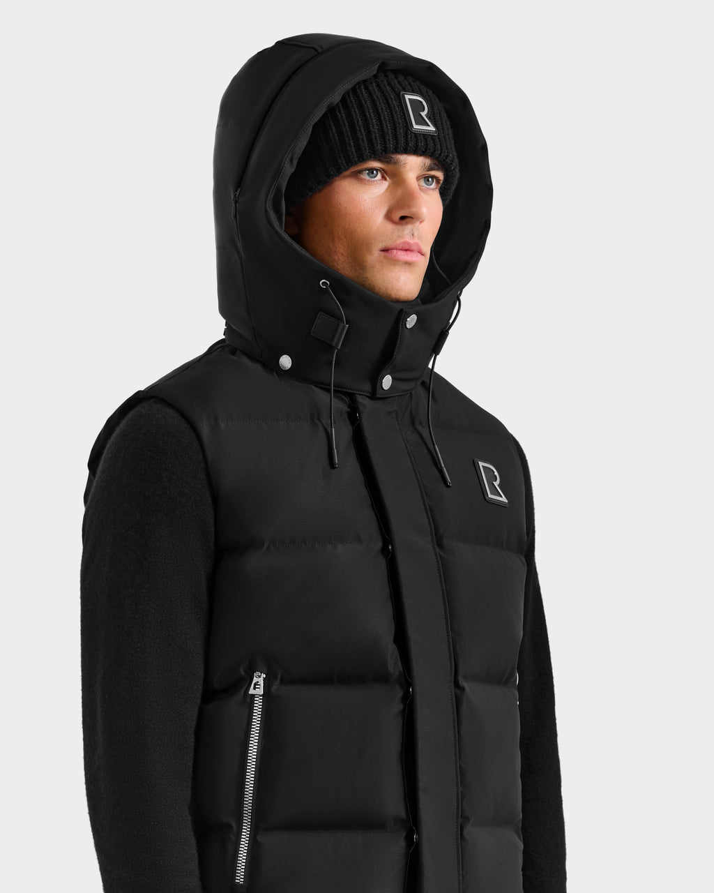 A person in the Rudsak REN H - 6125688 Black men’s quilted down puffer vest with removable hood and a matching black knit beanie, both featuring the Rudsak square R logo, stands against a plain light grey background—an essential cold-weather look.