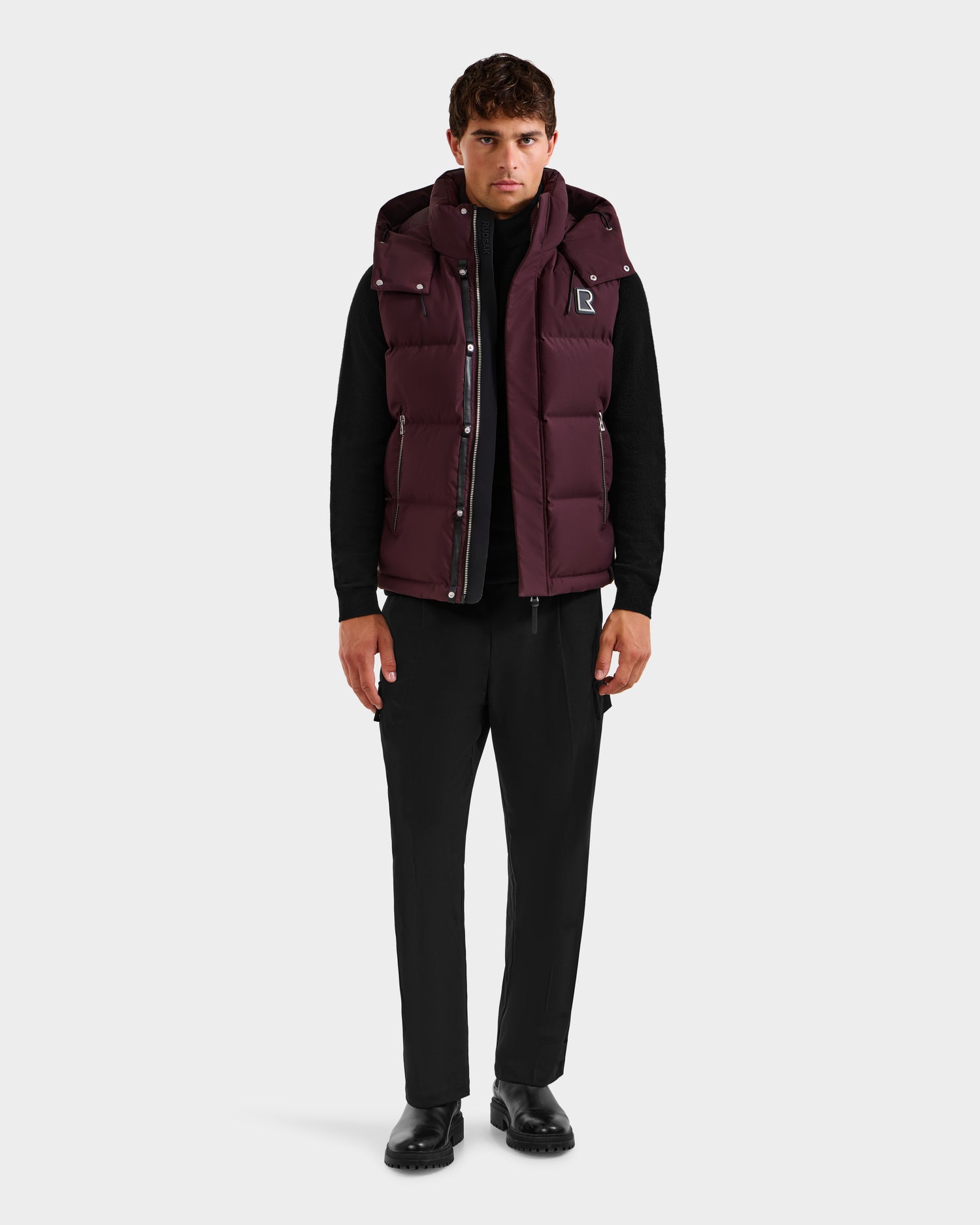A man faces forward, wearing the Rudsak REN H - 6125688 Bordeaux men’s quilted down puffer vest with removable hood over a black long-sleeve shirt, black pants, and black shoes, set against a plain white background.