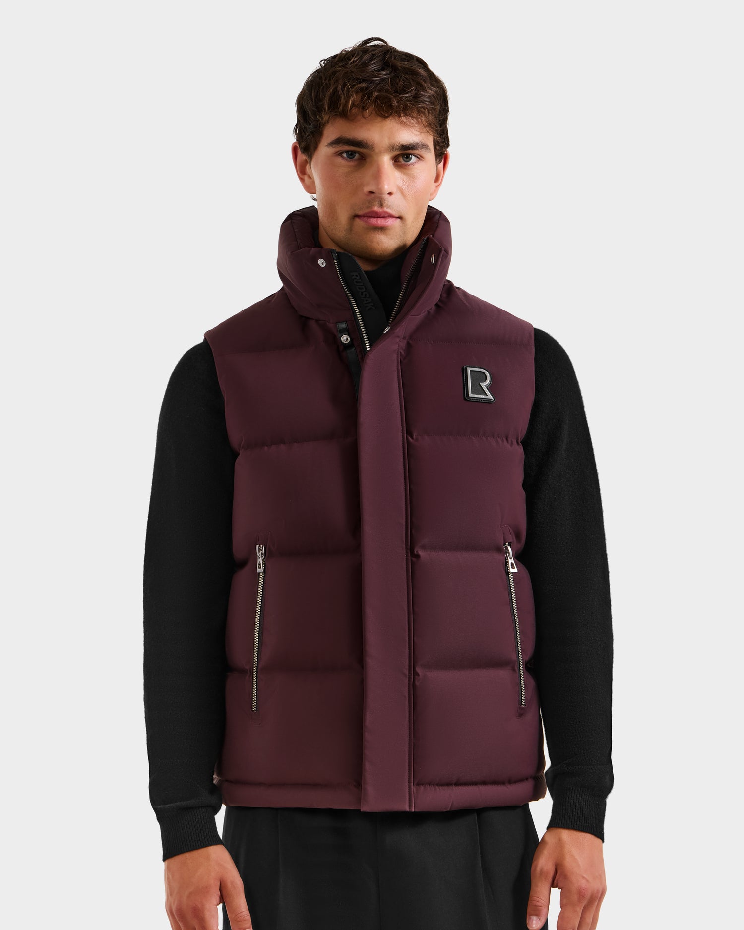 A man wearing the Rudsak REN H - 6125688 Bordeaux men’s quilted down puffer vest with zippered pockets, high collar, removable hood, and square chest logo over a black long-sleeve shirt stands against a white background.