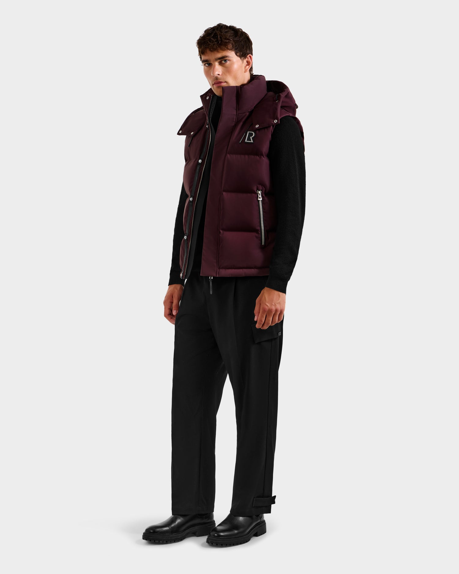 A man wears the Rudsak REN H - 6125688 Bordeaux men’s quilted down puffer vest with removable hood, styled over black basics—highlighting this cold-weather essential against a plain light grey background.