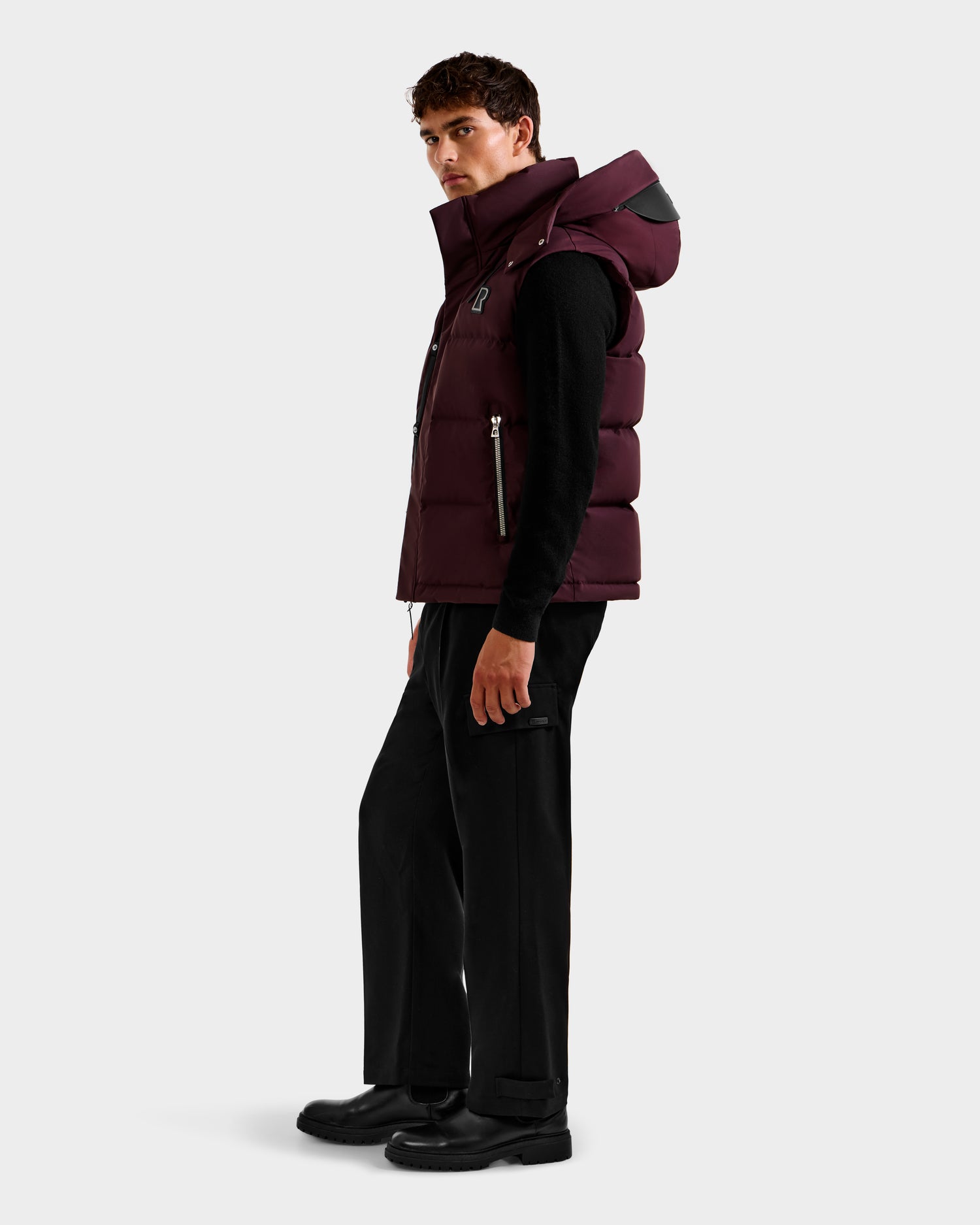 A man stands sideways wearing the Rudsak REN H - 6125688 Bordeaux quilted down puffer vest with a high collar and removable hood, over a black shirt and pants, against a plain light gray background.