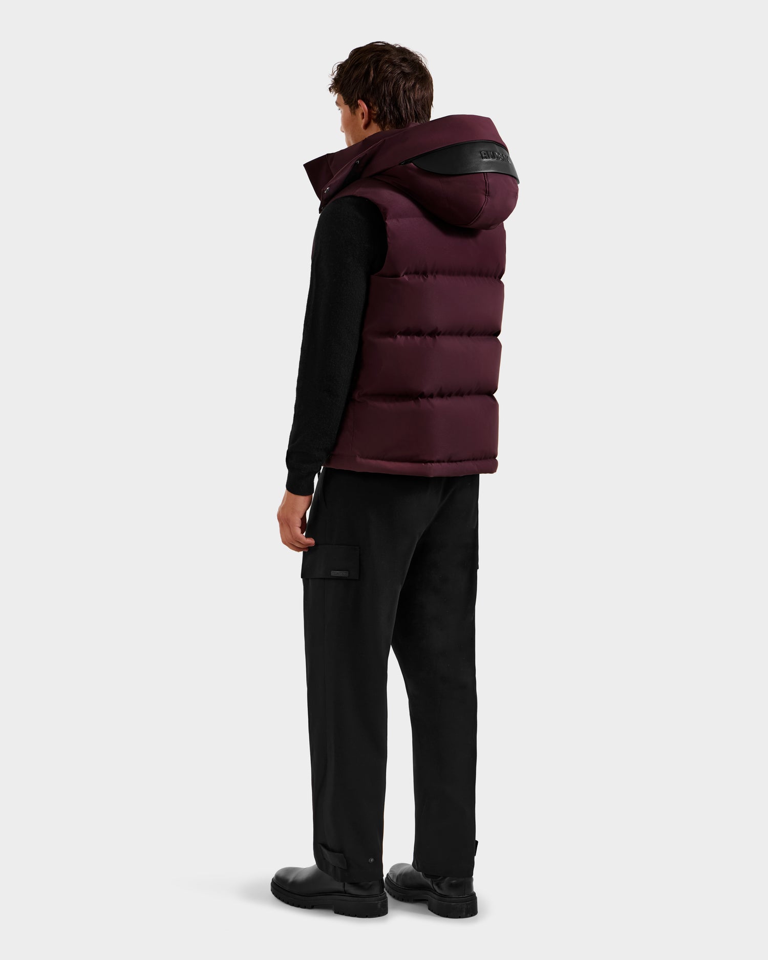 A person faces away, wearing the Rudsak REN H - 6125688 Bordeaux men’s quilted down puffer vest with removable hood, black long-sleeve shirt, cargo pants, and shoes—styled as a cold-weather essential against a white background.