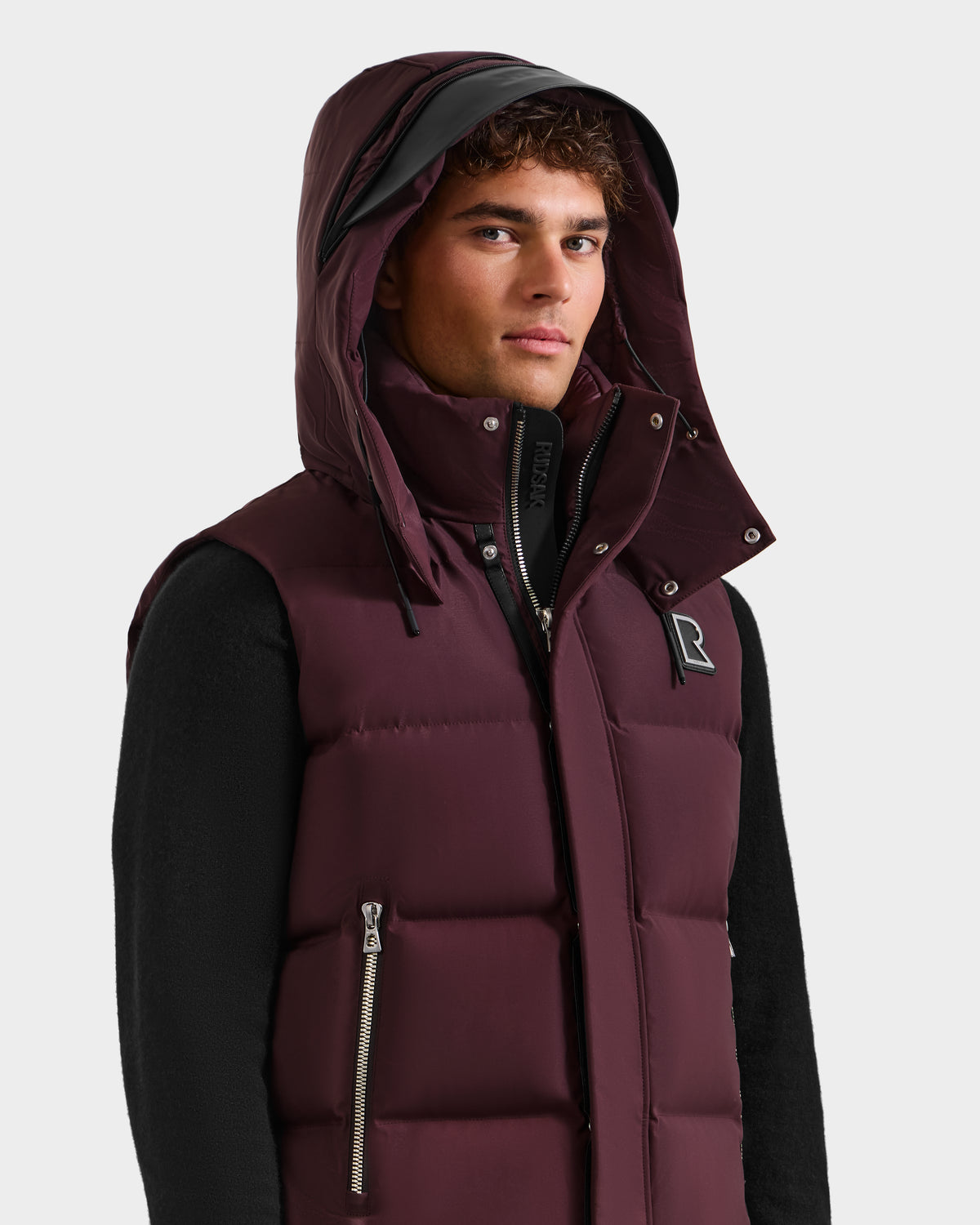 A young man wears the REN H - 6125688 Bordeaux quilted down puffer vest by Rudsak, featuring a removable hood, over a black long-sleeve shirt. He stands against a plain light background, looking at the camera—a true cold-weather must-have.