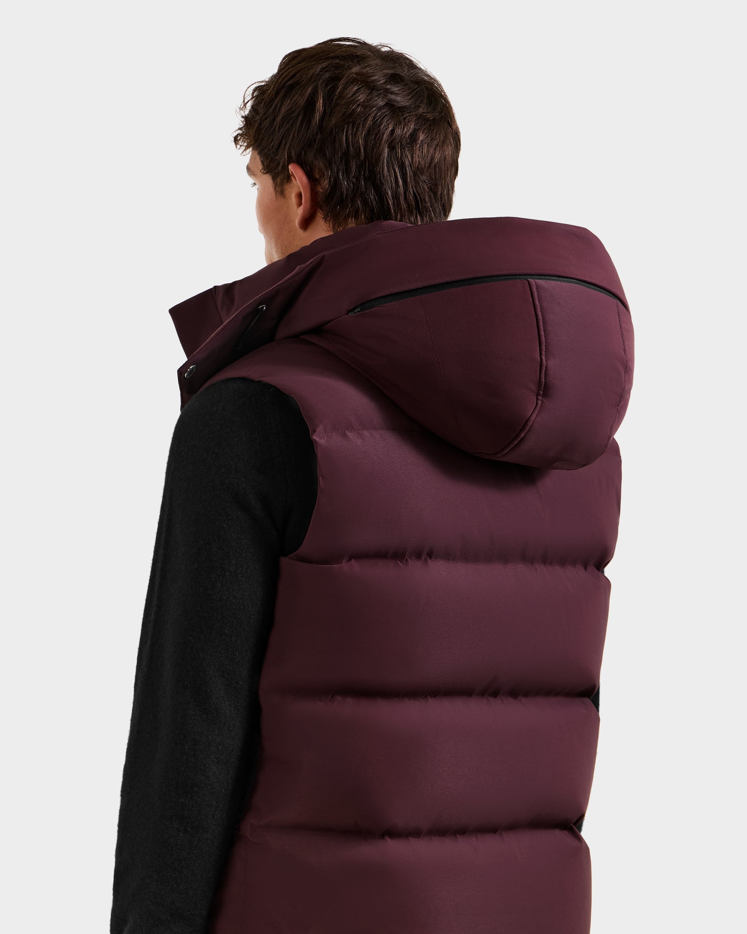A person in the Rudsak REN H - 6125688 Bordeaux men’s quilted down puffer vest with removable hood, layered over a black long-sleeve shirt, is shown from the back, slightly turned left. The background is plain and light-colored.