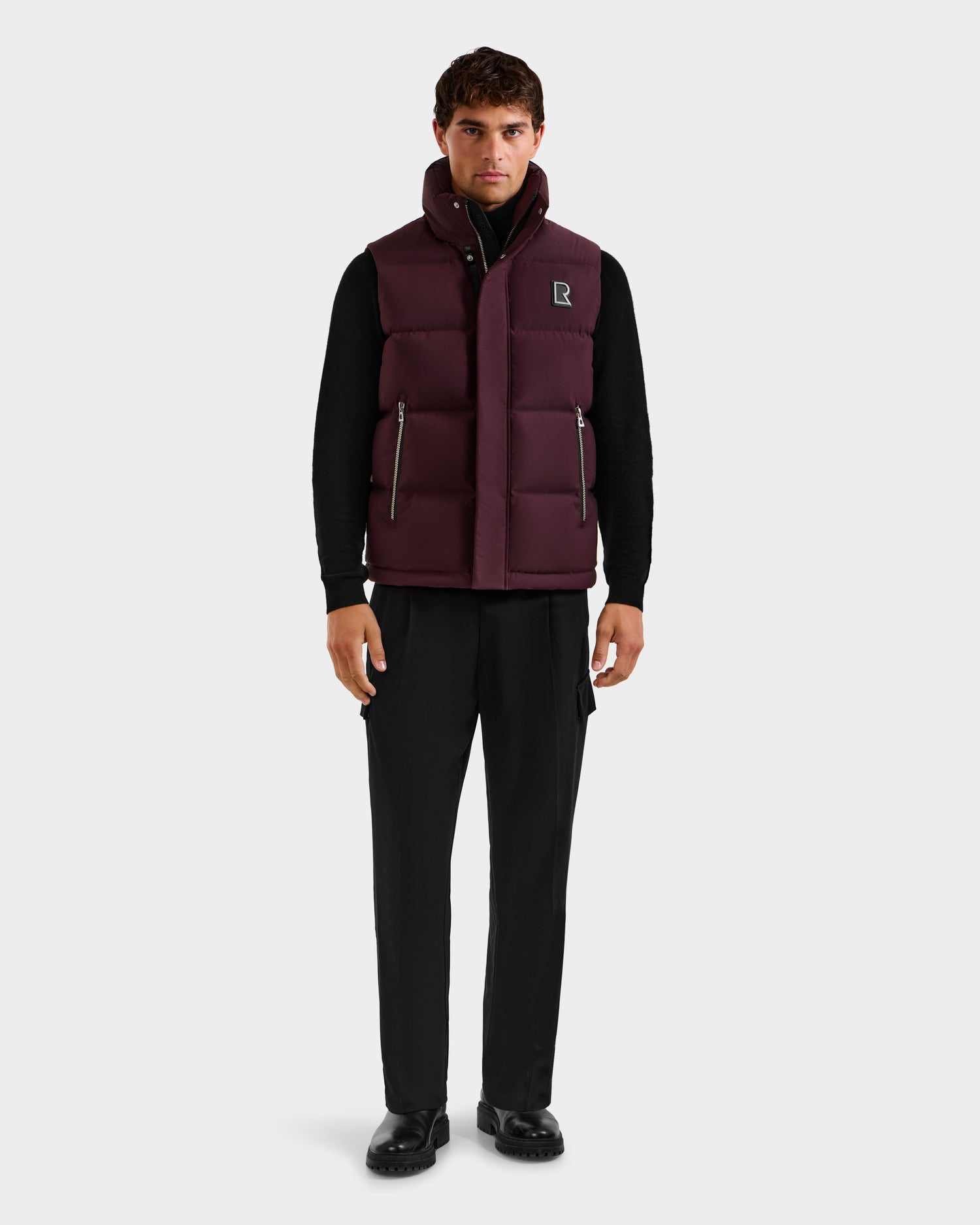 A man faces forward in a Rudsak REN H - 6125688 Bordeaux quilted down puffer vest over a black long-sleeve shirt, black pants, and black shoes. The background is plain white.