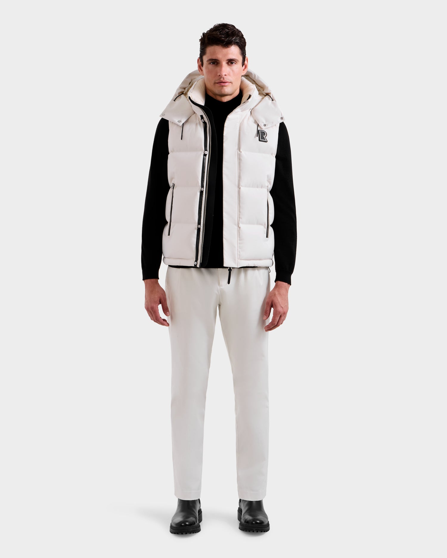 A man faces forward wearing the Rudsak REN H - 6125688 Vanilla men’s quilted down puffer vest with removable hood, paired with a black shirt, white pants, and black boots against a light background—a cold-weather essential.