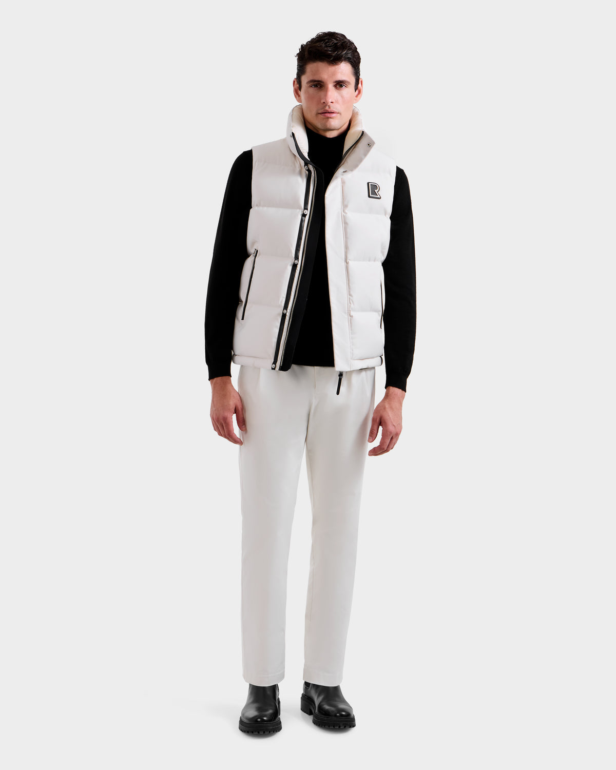 A man stands facing forward, wearing the Rudsak REN H - 6125688 Vanilla men’s quilted down puffer vest with a removable hood, over a black long-sleeve shirt, white pants, and black shoes against a plain light background.