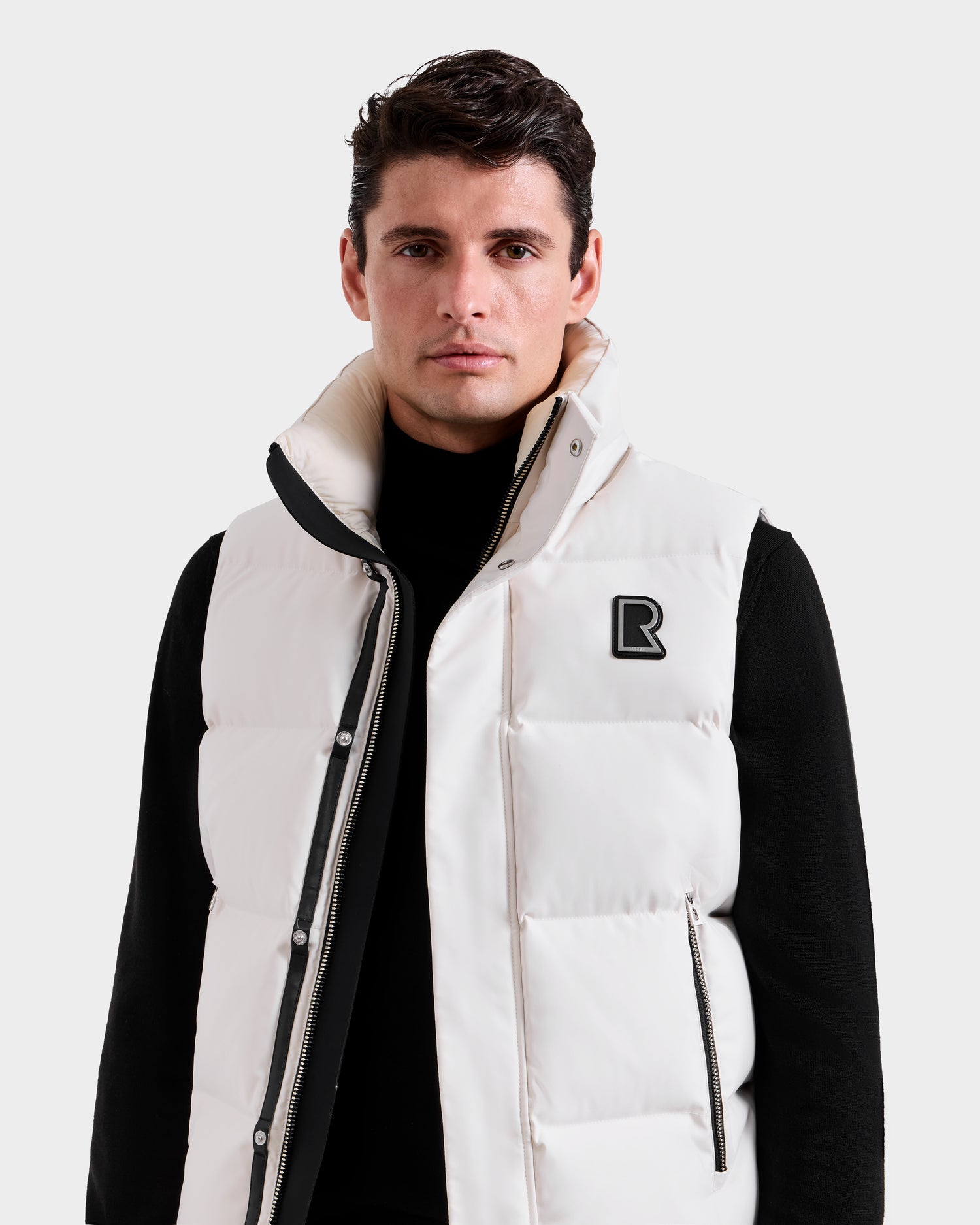 A man with short dark hair wears the Rudsak REN H - 6125688 Vanilla men’s quilted down puffer vest over a black turtleneck sweater, standing against a plain light gray background—a cold-weather essential for any modern wardrobe.