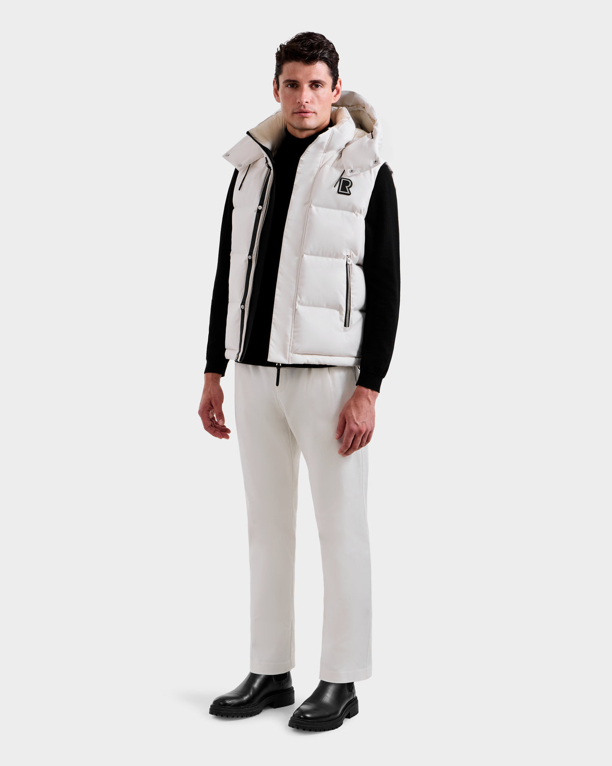 A man faces forward, wearing the Rudsak REN H - 6125688 Vanilla quilted down puffer vest with removable hood over a black long-sleeve top, white pants, and black boots against a light gray background—a cold-weather essential.