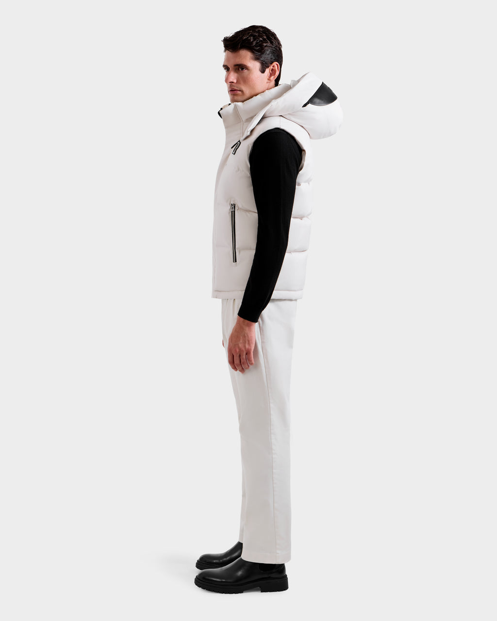 A man stands in profile against a plain background, wearing the Rudsak REN H - 6125688 Vanilla men’s quilted down puffer vest with removable hood, a black long-sleeve shirt, white pants, and black boots—an ideal cold-weather essential.