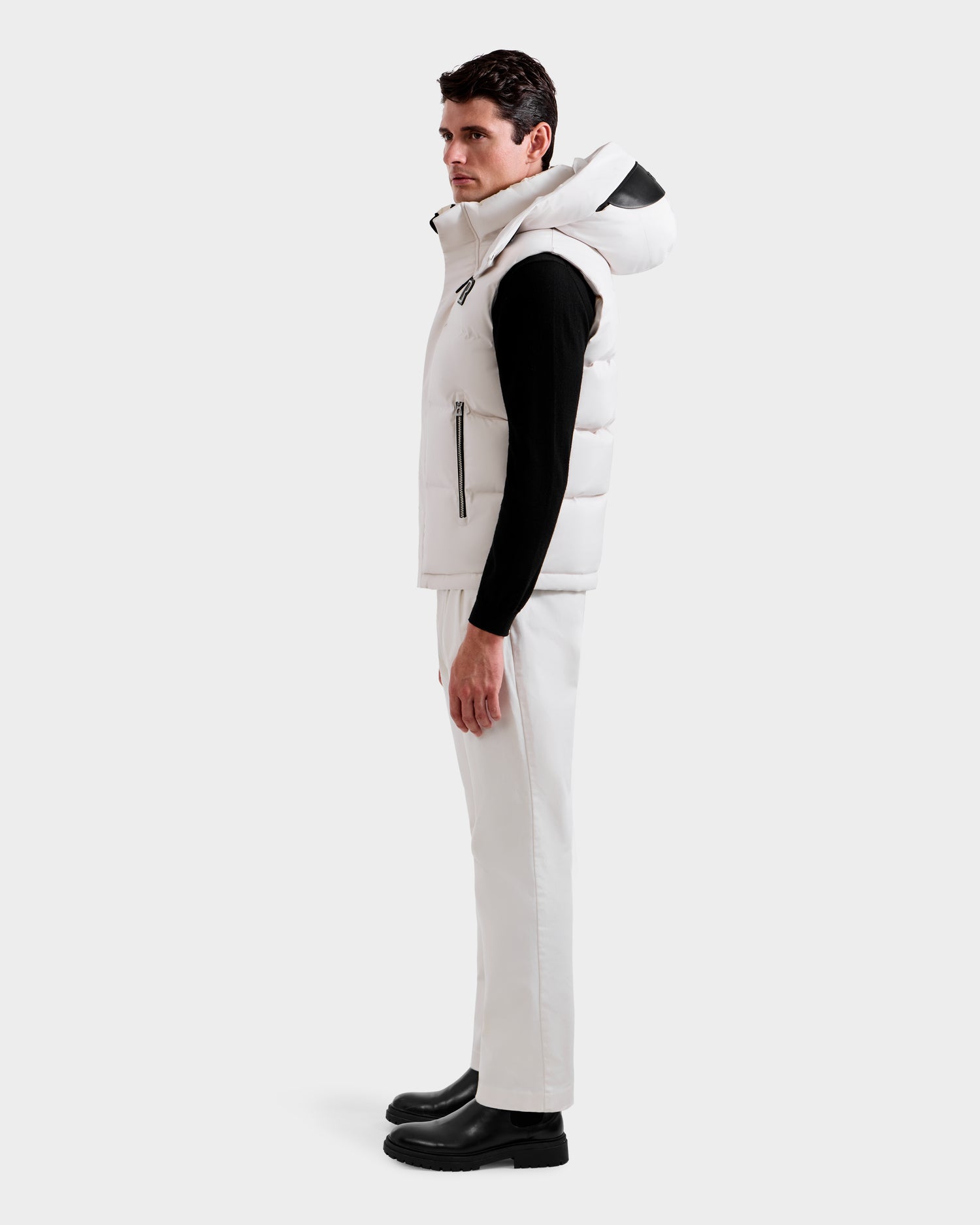 A man stands in profile against a plain background, wearing the Rudsak REN H - 6125688 Vanilla men’s quilted down puffer vest with removable hood, a black long-sleeve shirt, white pants, and black boots—an ideal cold-weather essential.