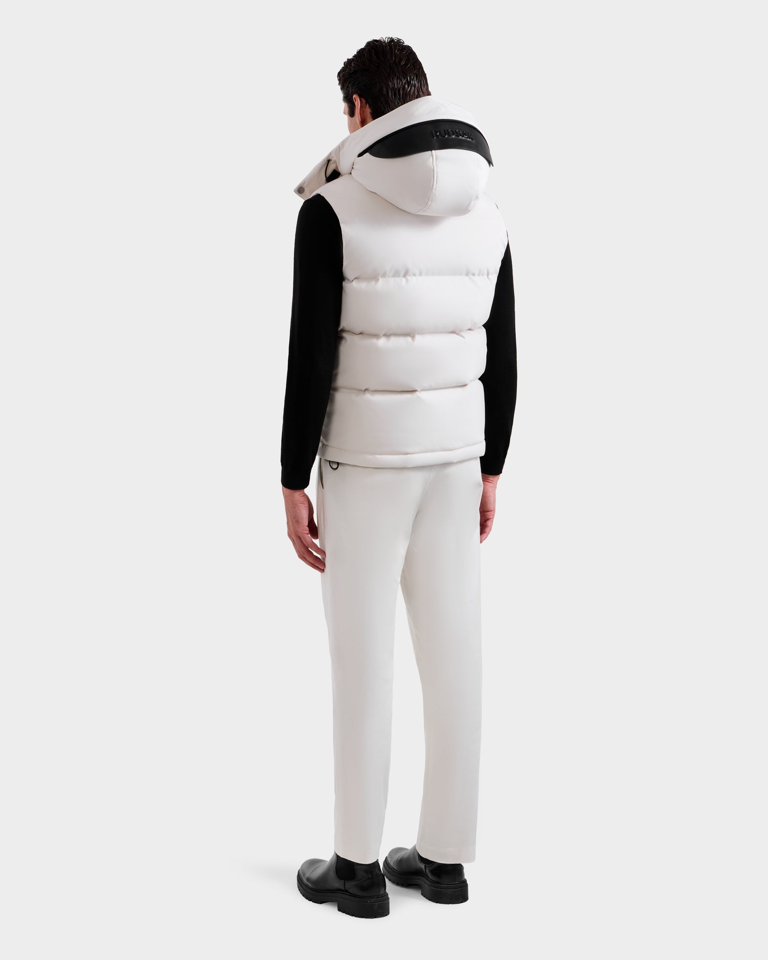 A man, facing away, wears the Rudsak REN H - 6125688 Vanilla men’s quilted down puffer vest with removable hood over a black long-sleeve shirt, white pants, and black shoes against a plain white background—a cold-weather must-have.