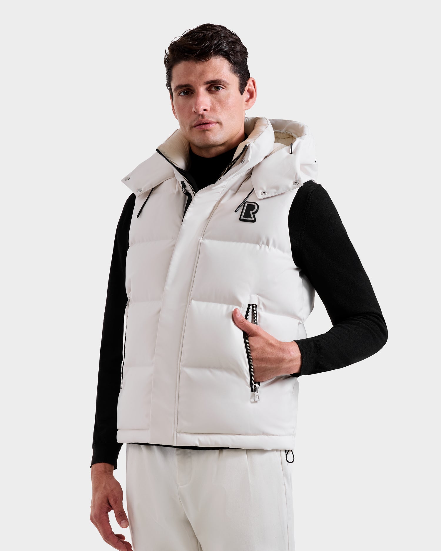 A man models the Rudsak REN H - 6125688 Vanilla quilted down puffer vest, featuring a removable hood and chest B logo, over a black turtleneck and white pants, set against a plain light background.