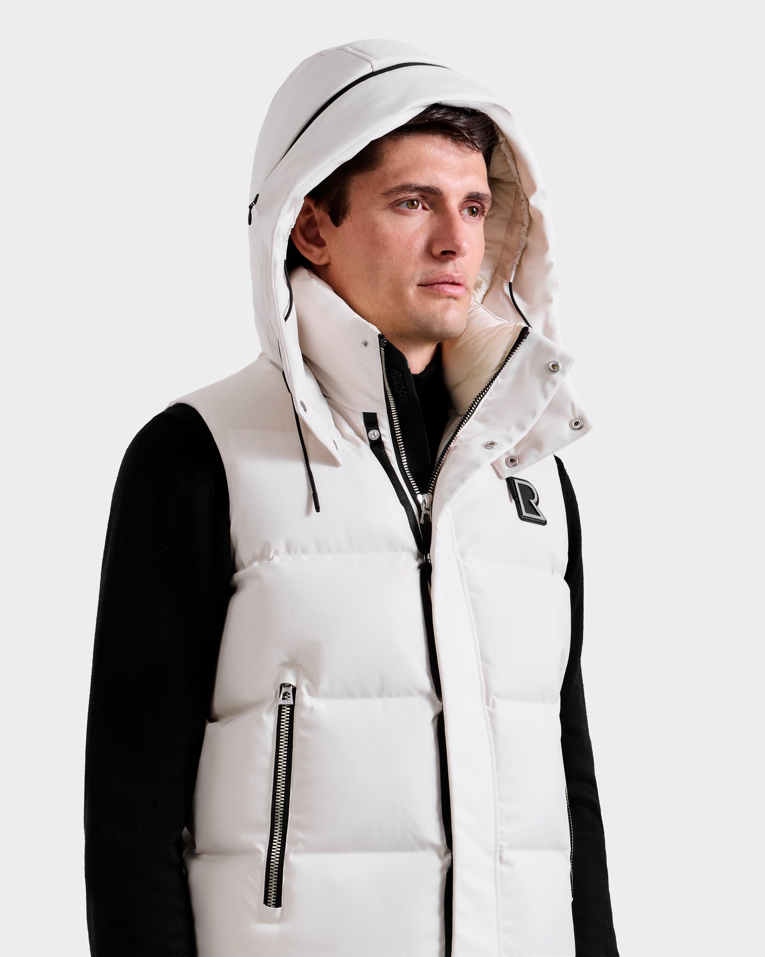 A man in the Rudsak REN H - 6125688 Vanilla quilted down puffer vest with a removable hood and zippered pockets, worn over a black long-sleeve shirt, stands against a plain light background. The vest features a chest R emblem.