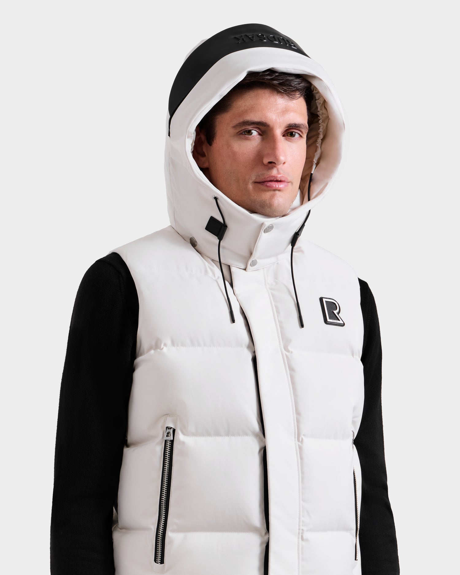 A man wears the Rudsak REN H - 6125688 Vanilla men’s quilted down puffer vest with removable hood and black accents over a black long-sleeve shirt. Features zippered pockets, embroidered R logo. Shown on a plain white background.