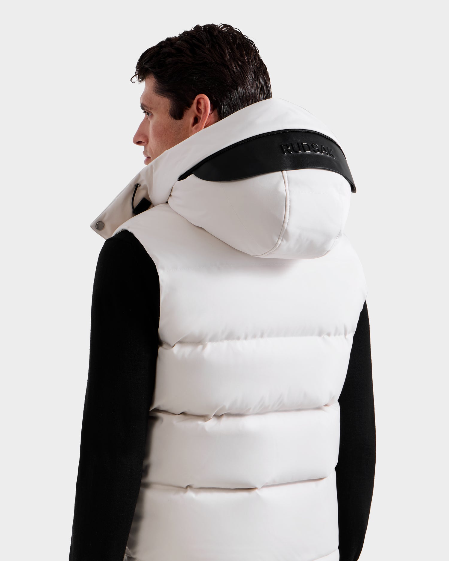 A man with dark hair, seen from behind, wears the Rudsak REN H - 6125688 Vanilla men’s quilted down puffer vest with black detailing and a large hood—partially showing the word MOOSE—over a black shirt, for warm and stylish comfort.