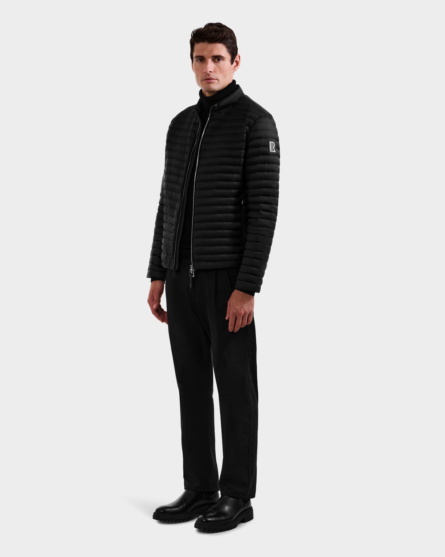 A man stands against a light gray background wearing the Rudsak REYMOND M - M125523 Black men’s down jacket, styled with black pants, a turtleneck, and shoes. He faces forward with one hand at his side and a neutral expression.