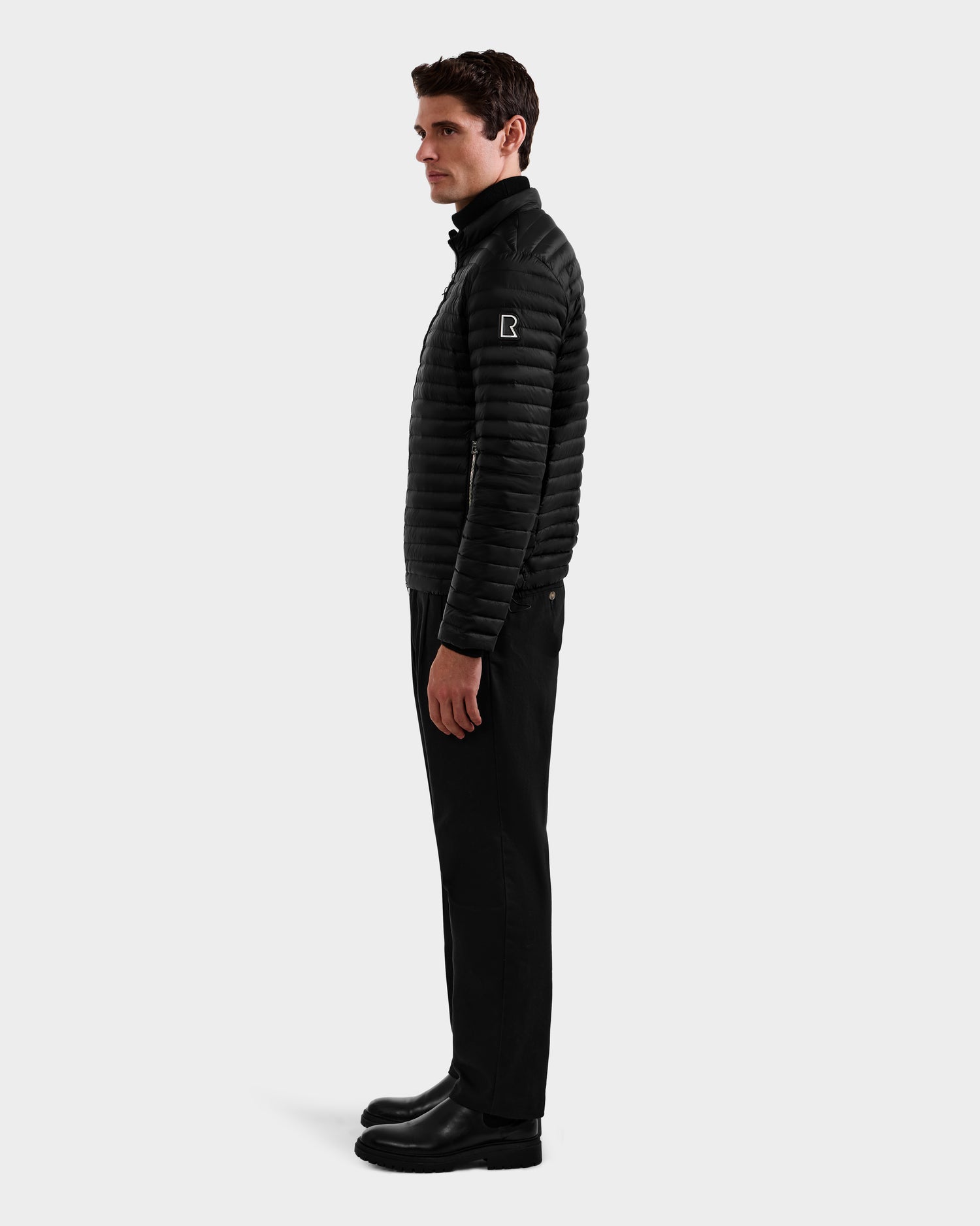 A man in profile facing left wears Rudsak's REYMOND M - M125523 Black quilted men’s down jacket, crafted from recycled nylon, paired with black pants and boots against a plain light gray background.